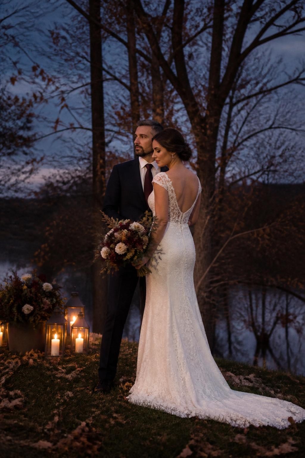 Realistic DSLR photo of a late fall wedding couple standing outdoors at twilight near bare trees and golden-brown leaves, bride holding a moody bouquet of burgundy, cream, and rust flowers, groom in a dark suit, faces softly turned away, lanterns glowing nearby, cool evening sky, cinematic romantic wedding style.