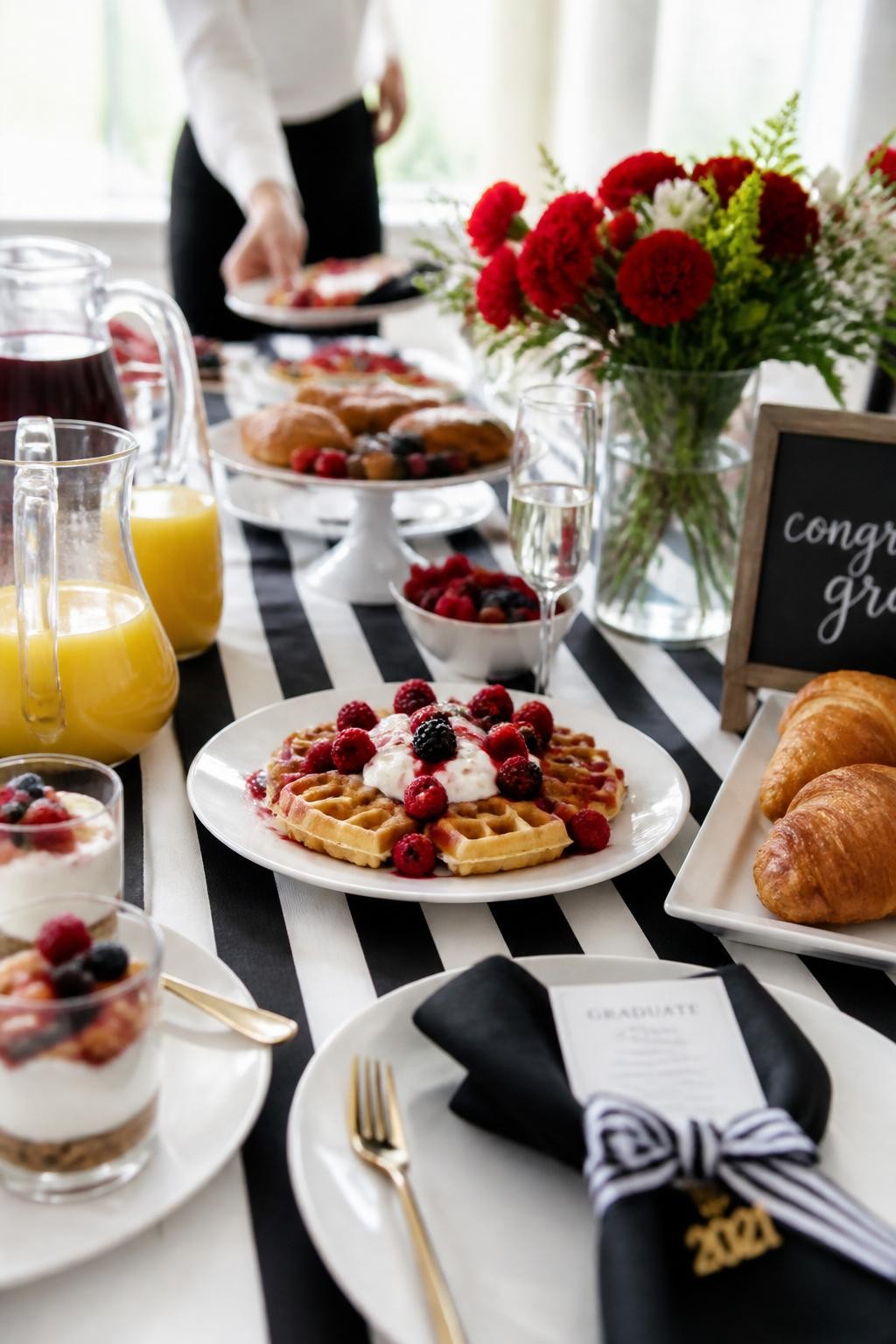 Image prompt: A realistic DSLR photo of a classy graduation brunch table with black and white striped details, red berry waffles, croissants, yogurt parfaits, glass juice pitchers, red flowers, black napkins, and one guest reaching for a plate in the background with face not visible and softly out of focus, bright morning window light, realistic brunch styling.