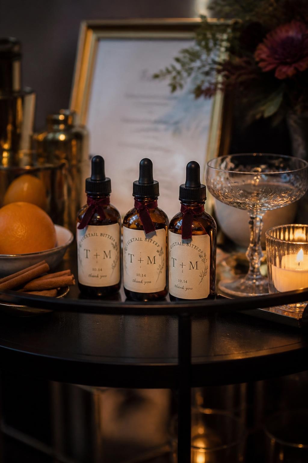 Realistic DSLR photo of tiny amber glass bottles of cocktail bitters with black dropper tops, custom cream labels, and velvet ribbon around the neck, styled on a dark bar cart with oranges, cinnamon sticks, coupe glasses, and candlelight, moody fall wedding cocktail hour setting.