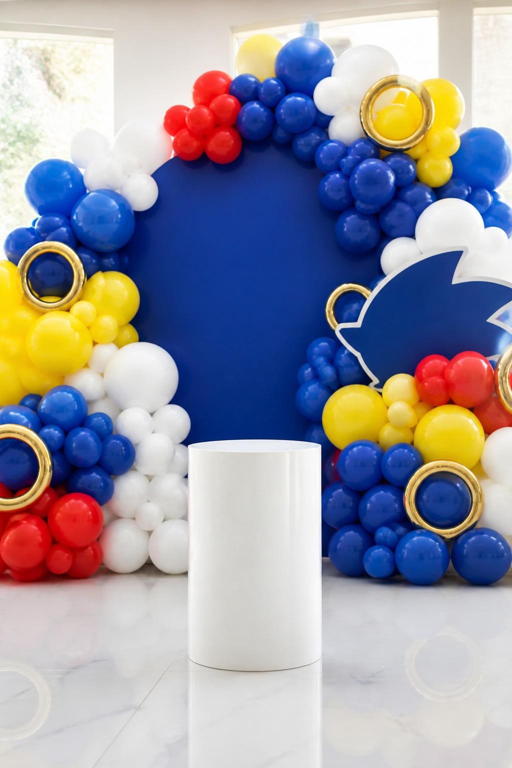 Image Prompt:
A single realistic photo of a modern Sonic birthday backdrop with a full balloon wall in royal blue, white, red, and yellow, arranged in different balloon sizes, small gold ring props tucked into the wall, clean white cake plinth in front, glossy indoor party floor, bright natural lighting, no people, no faces, realistic party photography, no text, no watermark.