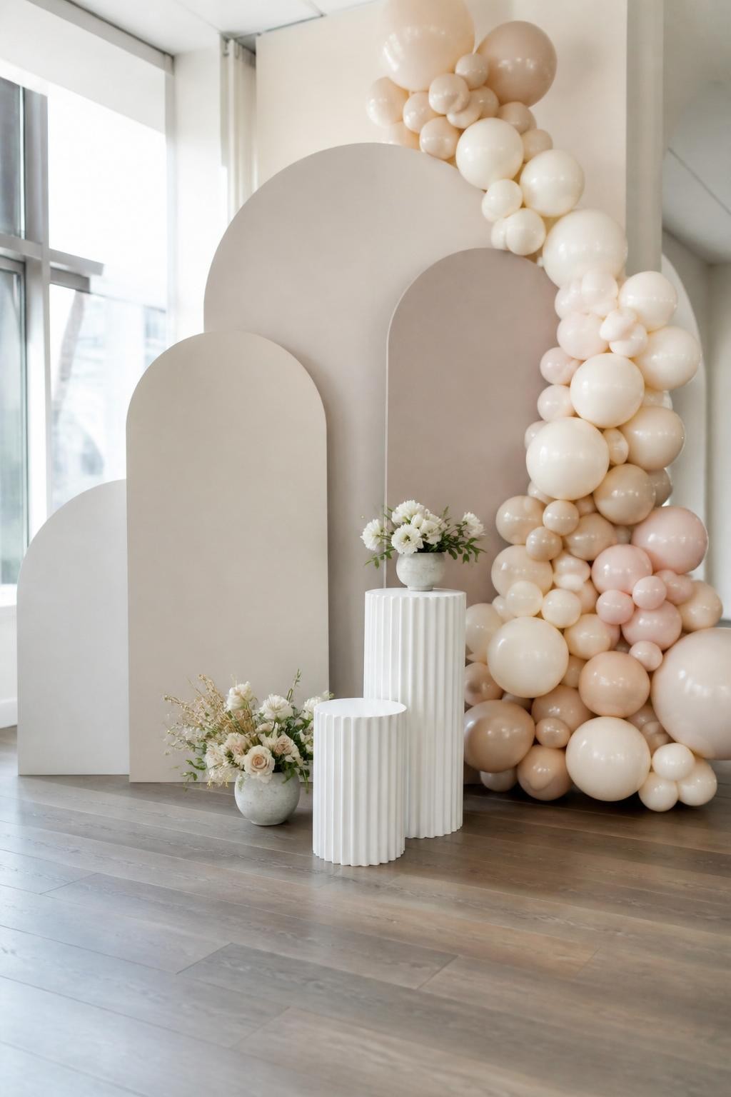 Image Prompt:
A single realistic editorial-style photo of a modern party arch backdrop with three layered curved arch panels in cream, beige, and soft taupe, arranged at different heights, organic balloon garland in ivory, champagne, and muted blush flowing down one side, small white plinths at the base, soft florals in low ceramic vases, clean indoor event space, no people, no faces, soft natural daylight, realistic DSLR event photography, no text, no watermark.