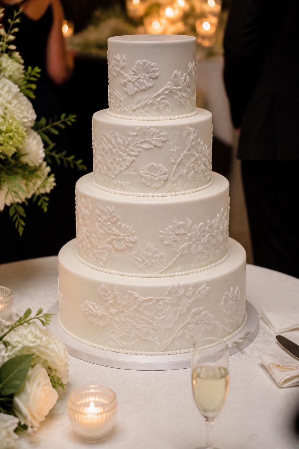 Image Prompt: Ultra-realistic wedding reception image featuring a white-on-white wedding cake with detailed bas-relief floral and vine patterns sculpted onto the tiers. Show the cake on a decorated cake table in a real wedding party scene with white flowers, candles, fine linens, and soft warm lighting. The cake should be the main focus. No visible faces. If any guests appear, show only cropped hands, soft back views, side silhouettes, or neck-down figures. Realistic raised icing detail, smooth finish, elegant and believable wedding styling, DSLR realism. (no watermarks on images)
