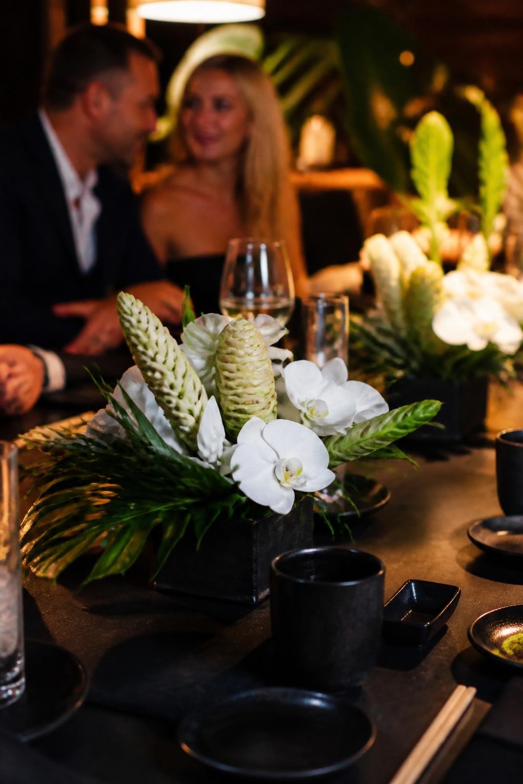 Image Prompt:
A realistic DSLR photo of a hibachi party dinner table with low centerpieces made of white orchids, ginger flowers, and lush greenery in compact modern vessels, black dinnerware, chopsticks, warm evening light, one couple seated out of focus in the background, elegant realistic event styling.