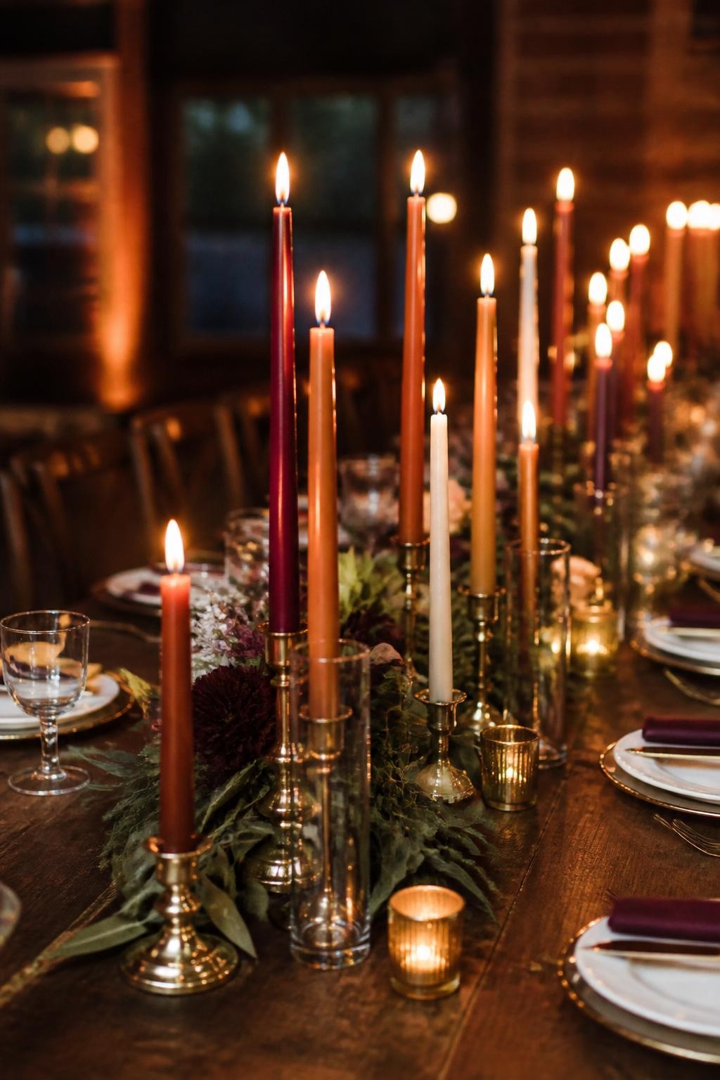 Realistic DSLR photo of a long fall wedding table decorated with mixed taper candles in burgundy, rust, mustard, plum, and ivory, placed in brass and glass holders at different heights, surrounded by low greenery, dark wood table, elegant plates, and warm candlelit reception atmosphere.