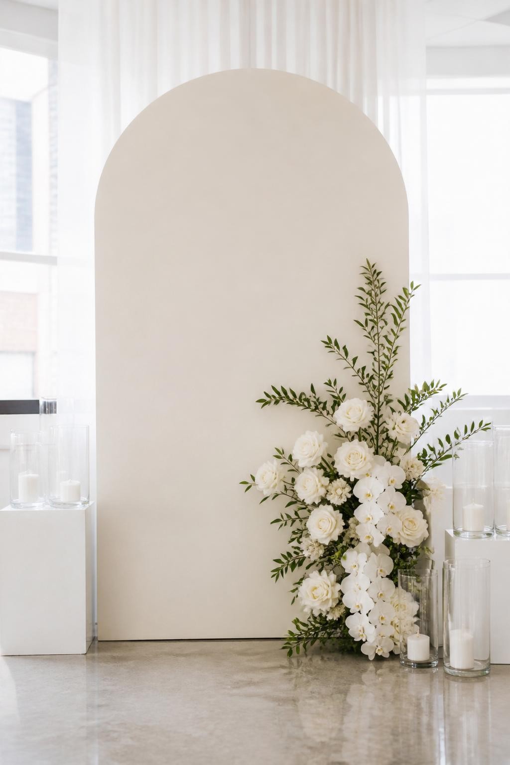 Image Prompt:
A single realistic editorial-style photo of a minimalist flower backdrop with one matte ivory arch panel, a small asymmetrical cluster of white roses, orchids, and greenery placed on the lower right side, clear candle holders, clean white plinths, bright indoor venue, no people, no faces, soft natural lighting, realistic upscale event photography, no text, no watermark.