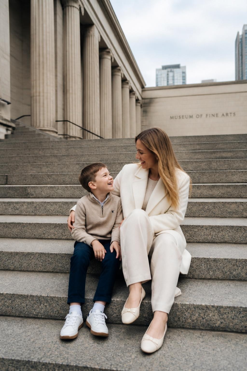 Image Prompt:
A realistic editorial mother and son photoshoot on the wide stone steps outside a modern art museum, featuring a white mother in a tailored cream coat, straight-leg trousers, and simple flats, sitting beside her young son in a soft beige sweater, navy trousers, and clean sneakers. They are seated close together, the son leaning gently against his mother while they look at each other and laugh naturally. The background has tall museum columns, clean stone textures, soft cloudy daylight, no crowd, calm city atmosphere, DSLR photo-realistic style, vertical image.