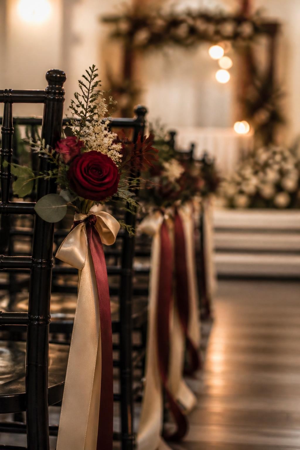 Realistic DSLR close-up of fall wedding aisle chairs decorated with small burgundy rose posies, cream filler flowers, dark greenery, and trailing silk ribbons in champagne and wine tones, tied to black wooden chairs, soft indoor ceremony light, blurred floral altar in the background, refined romantic style.
