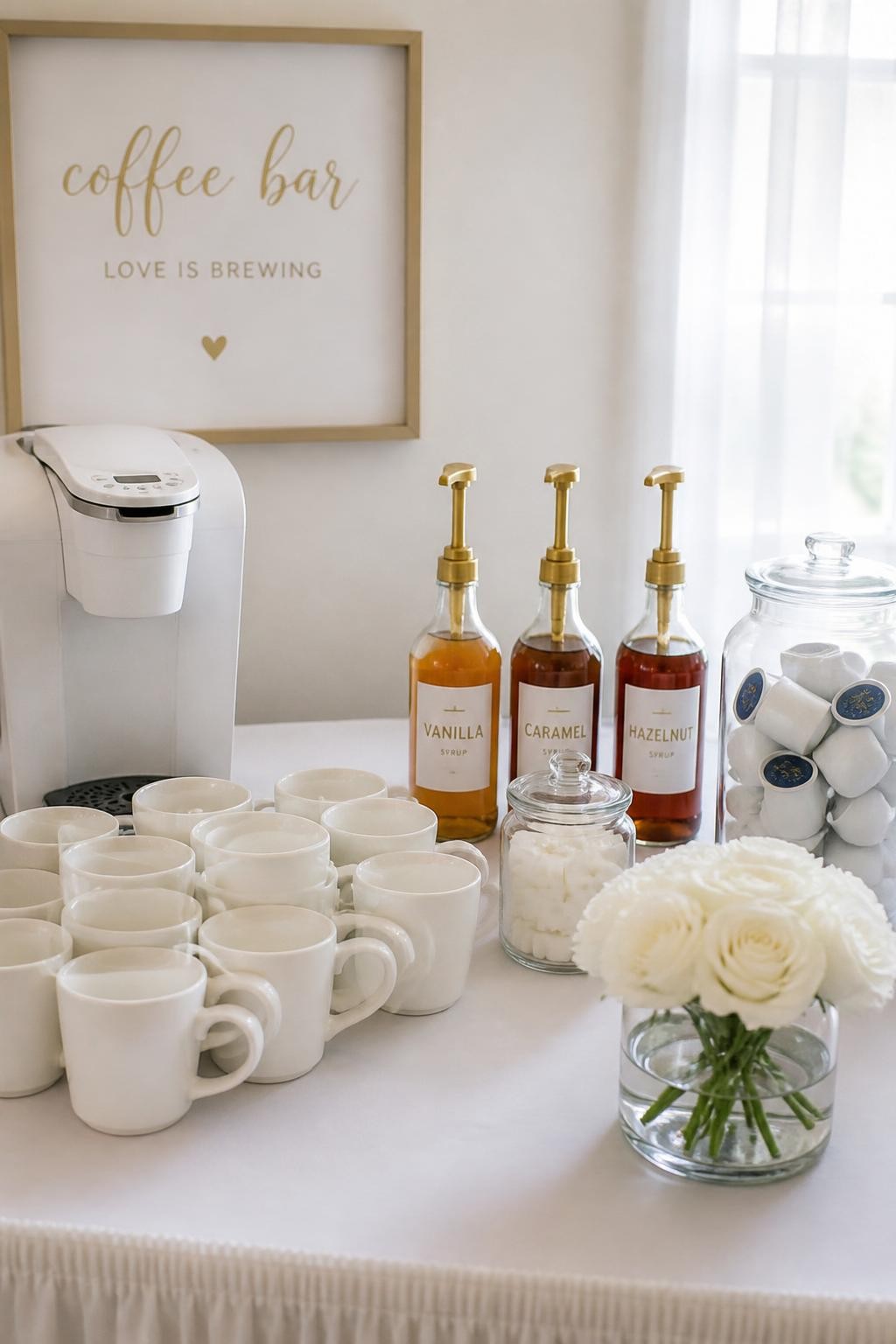 A realistic DSLR photo of a minimalist bridal shower coffee bar, clean white table, white coffee machine, white mugs stacked in rows, glass bottles with gold labels for vanilla, caramel, and hazelnut syrup, clear jars of sugar cubes and coffee pods, single low vase of white roses, soft daylight, elegant uncluttered party styling, no people.