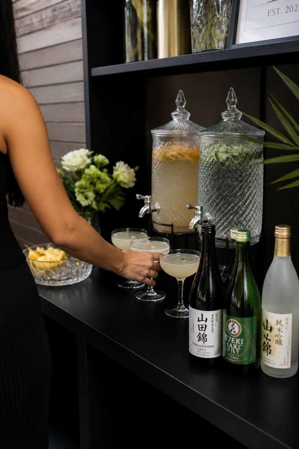 Image Prompt:
A realistic DSLR photo of a hibachi party drink display with sake bottles, clear mocktail dispensers, coupe glasses, citrus garnishes, black shelving, soft lighting, and one guest reaching for a drink with face out of frame, chic entertaining photography.