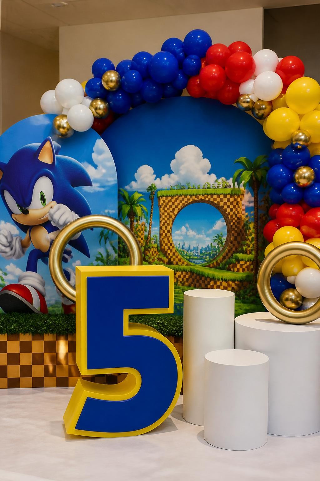 Image Prompt:
A single realistic editorial-style photo of a Sonic-inspired birthday backdrop with a large freestanding birthday age number prop in royal blue and yellow, bright balloon garland in blue, red, white, and yellow, gold ring props, checkered platform accents, white plinths, indoor party setup, no people, no faces, realistic event photography, no readable text, no watermark.