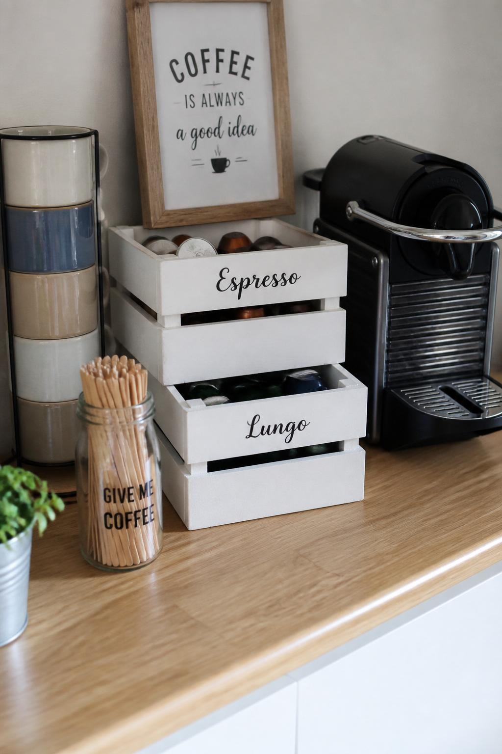 A realistic DSLR photo of a small coffee bar with painted mini wooden crates used as coffee pod storage, two stacked white crates holding colorful coffee pods, compact coffee machine beside them, ceramic mug stack, small framed coffee sign, glass jar of stirrers, light wood countertop, warm natural light, realistic DIY home styling, no people.