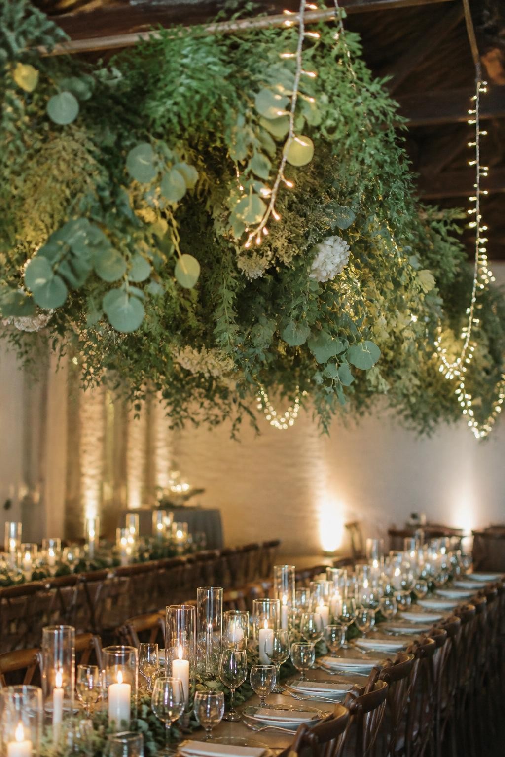 Detailed image prompt: A realistic photo of hanging greenery garlands suspended above wedding reception tables, lush green vines, eucalyptus, ruscus, and small white flowers, warm fairy lights woven through, long tables underneath with candles and glassware, romantic indoor wedding venue, no people.