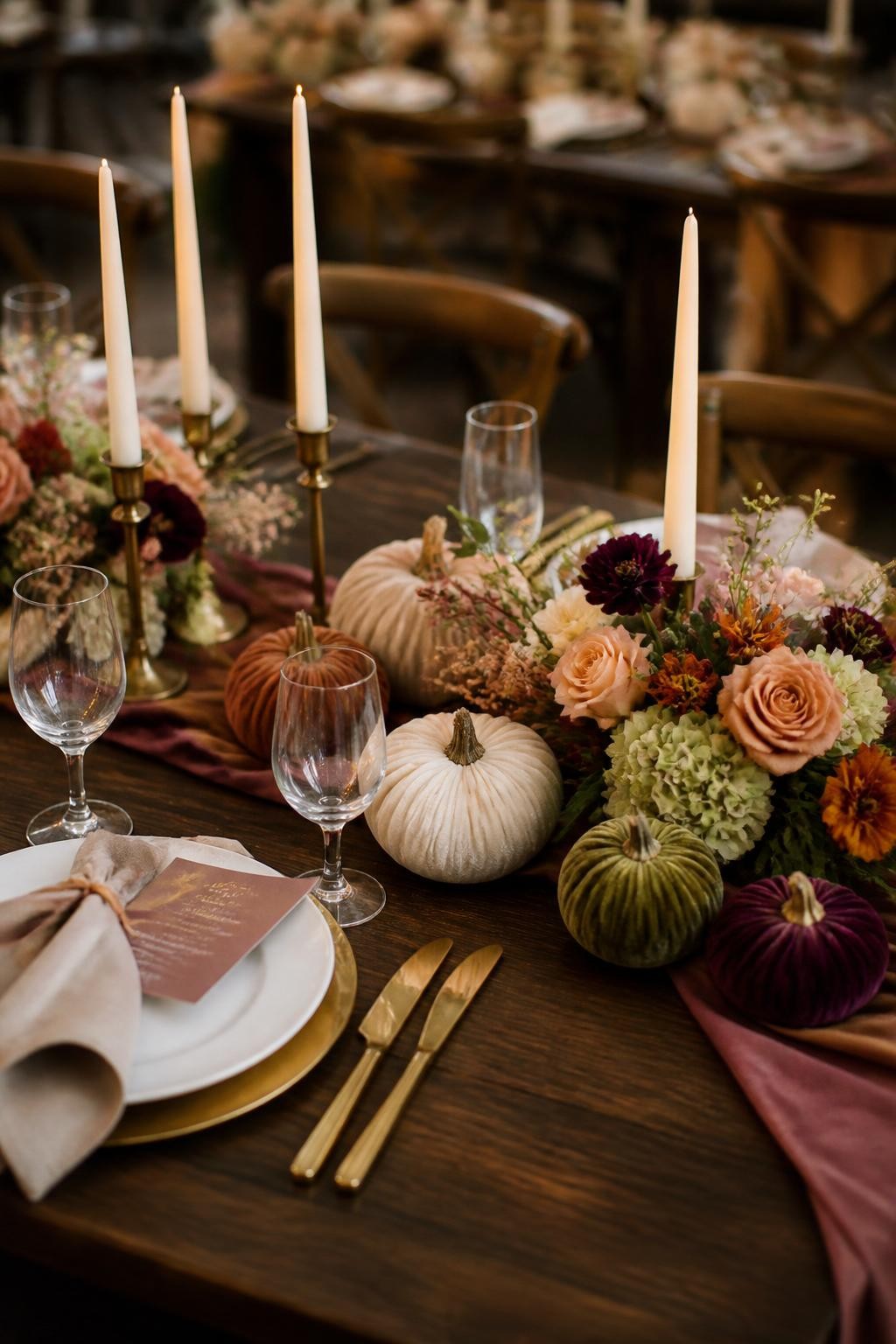 Realistic DSLR photo of a luxury fall wedding table centerpiece featuring velvet pumpkins in champagne, rust, olive, and plum, surrounded by low floral sprays, brass candle holders, ivory taper candles, and silk ribbon details, set on a dark wood table with elegant place settings, warm indoor reception lighting.