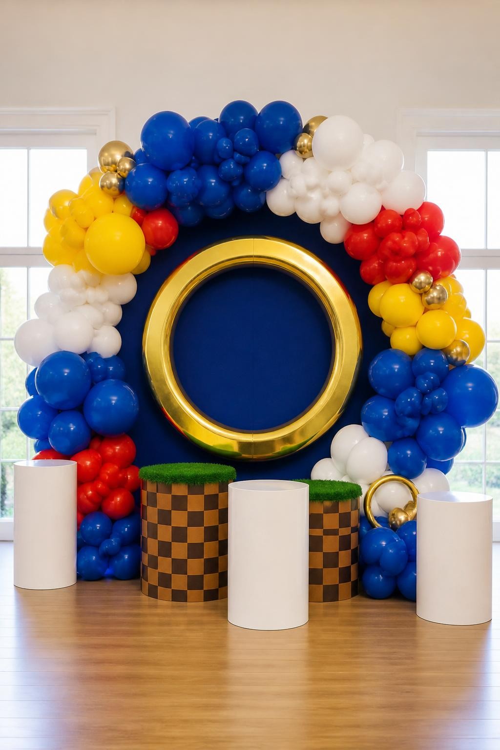 Image Prompt:
A single realistic editorial photo of a Sonic party backdrop with a circular balloon ring arch made of royal blue, white, yellow, and red balloons, large gold ring prop in the center, small checkered platform details at the base, white plinths, bright indoor birthday venue, no people, no faces, realistic high-quality event photography, no text, no watermark.