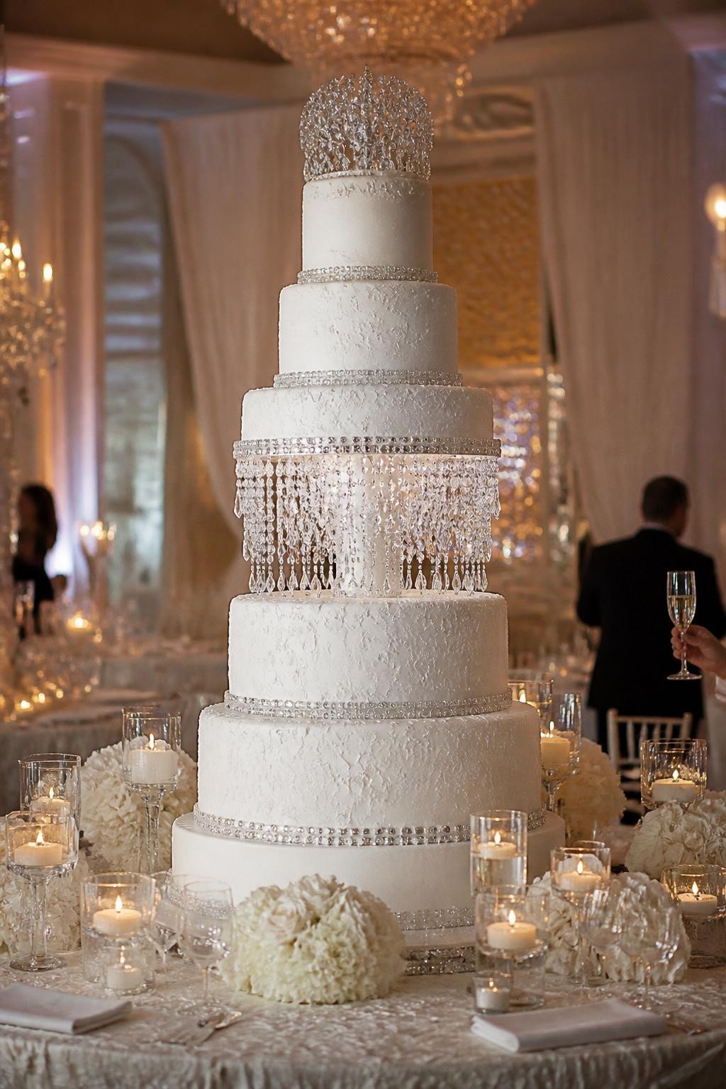 Image Prompt: Ultra-realistic wedding reception photo of an extravagant crystal chandelier wedding cake with tall white tiers, crystal bands, and hanging crystal details that look like a chandelier. Place the cake on a luxury cake table in a real wedding party scene with candles, white flowers, elegant linens, crystal glassware, and a grand ballroom backdrop. Keep the cake as the main focus. No visible faces. If people appear, show only blurred back views, soft side views, cropped-from-neck figures, or hands holding champagne glasses. Realistic icing texture, believable crystal sparkle, romantic lighting, DSLR editorial wedding photography. (no watermarks on images)
