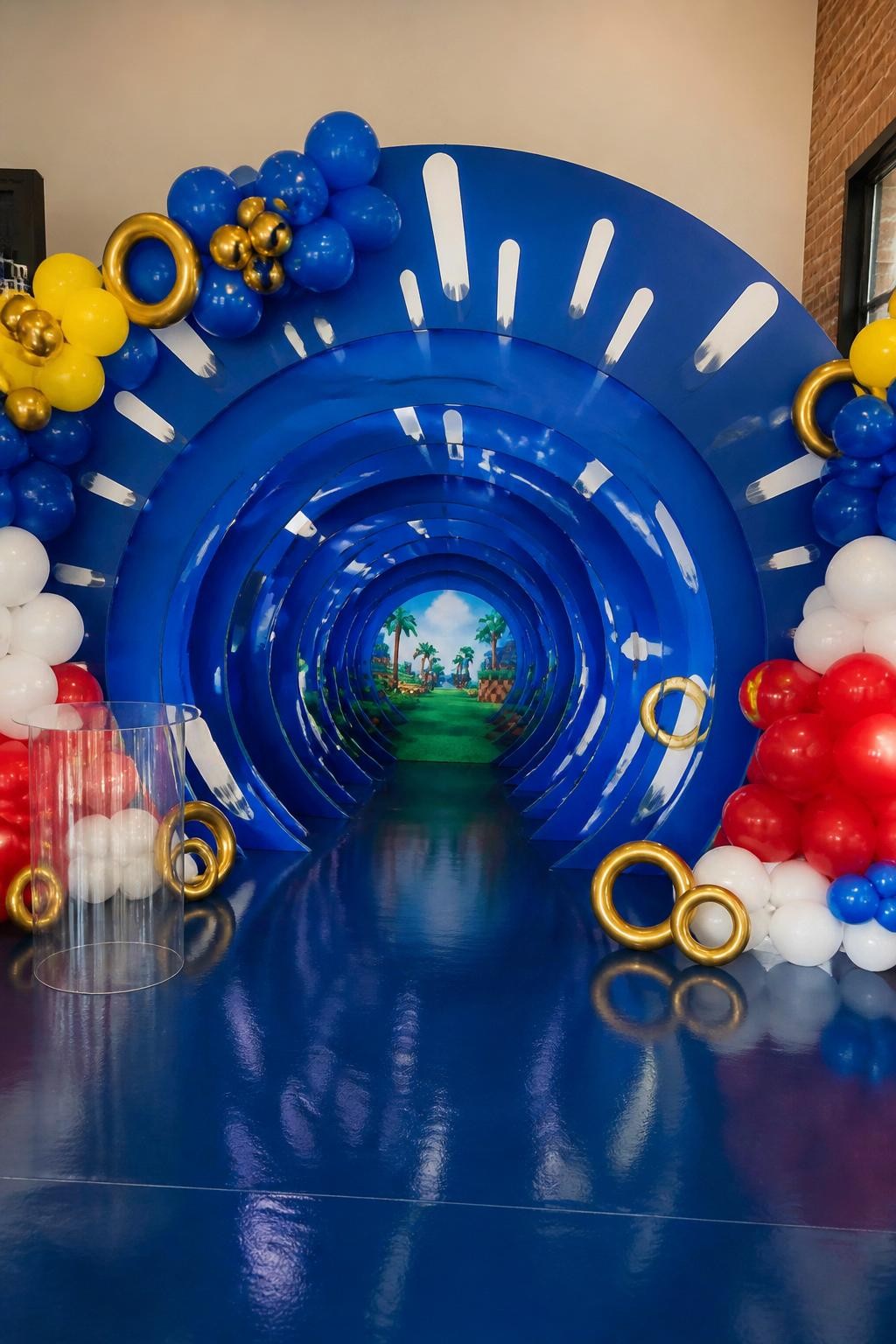 Image Prompt:
A single realistic photo of a Sonic-themed party backdrop designed like a speed tunnel, with layered circular blue panels, white motion streak accents, yellow balloon details, red and white balloon clusters, a glossy floor, small gold ring props, indoor birthday setup, no people, no faces, realistic energetic event photography, no readable text, no watermark.