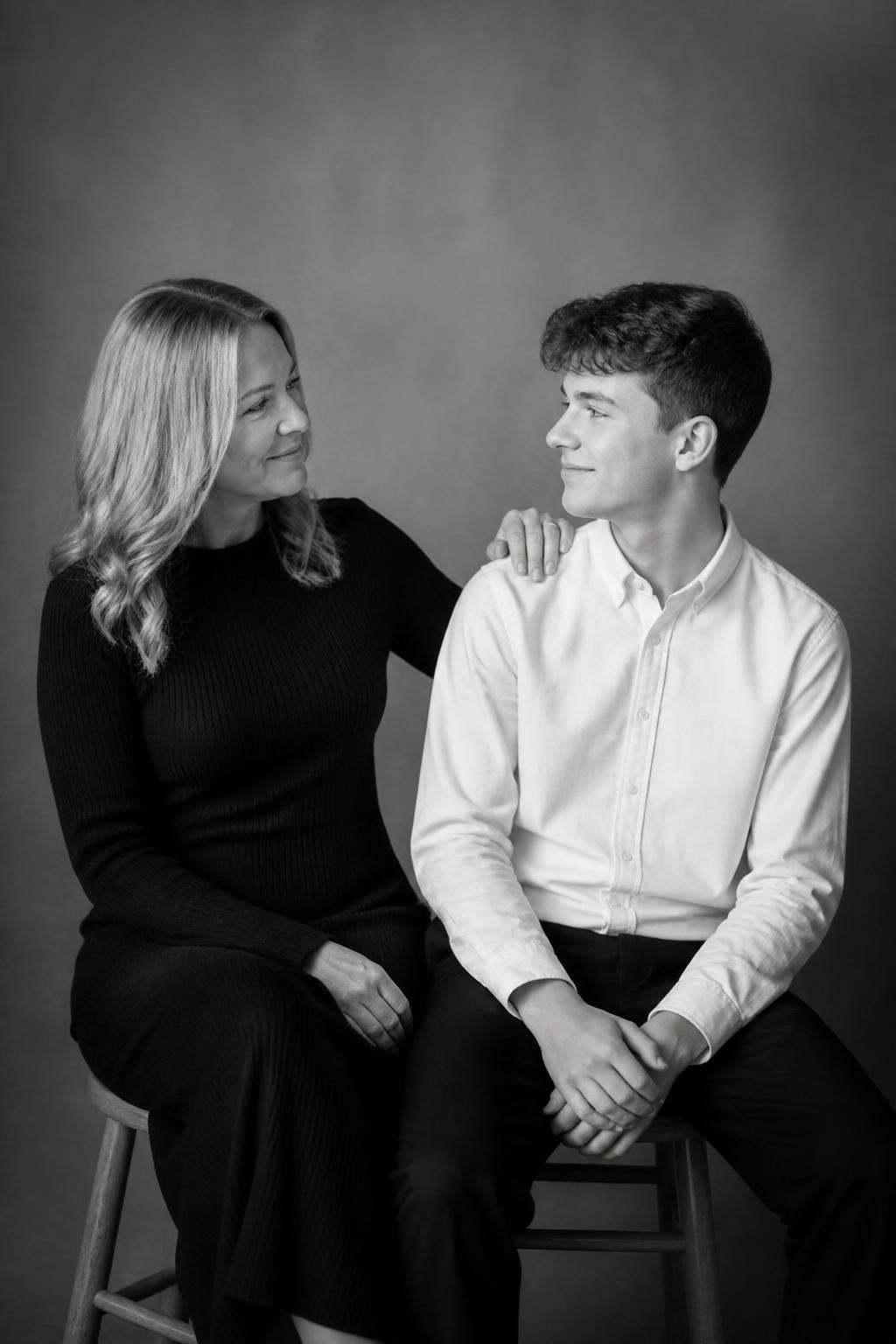 Image Prompt:
A realistic black-and-white studio portrait of a white mother and her teenage son seated on simple wooden stools against a warm gray seamless backdrop. The mother wears a long-sleeve black knit dress, and the son wears a white button-up shirt with dark trousers. They sit close but not overly posed, with the mother’s hand resting lightly on her son’s shoulder while he looks toward her with a small smile. Soft directional studio light, gentle shadows, timeless heirloom portrait style, high-resolution DSLR photography, vertical composition.