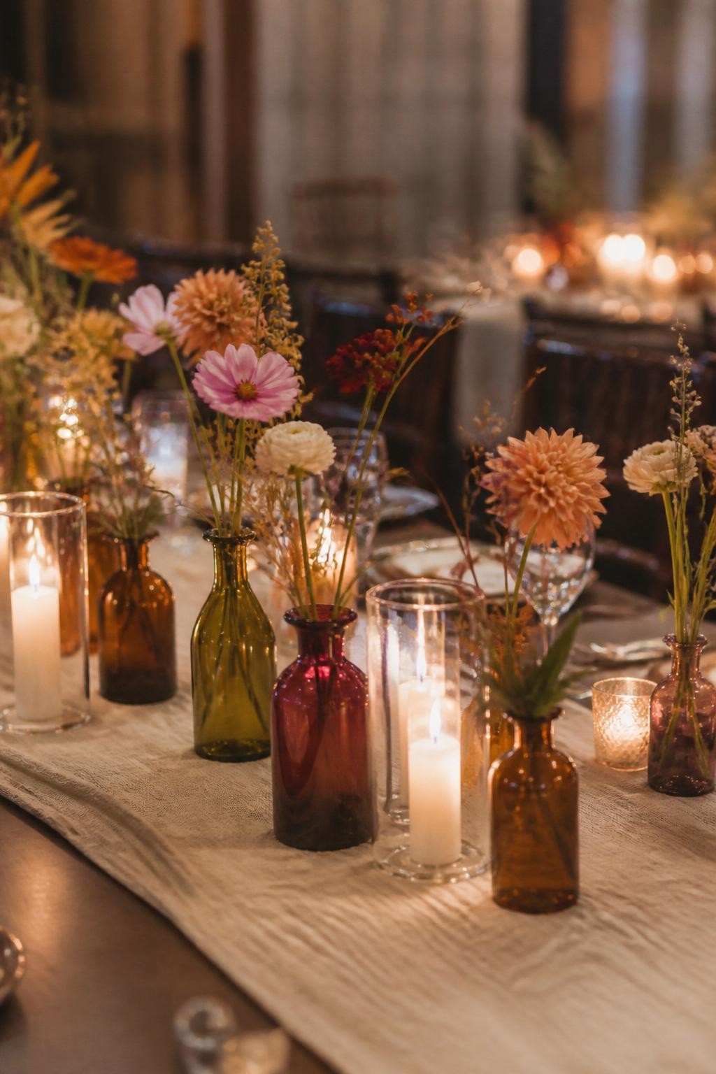 Realistic DSLR photo of a fall wedding table decorated with small colored glass bud vases in amber, cranberry, olive, and smoky purple, each holding single stems of dahlias, ranunculus, cosmos, dried grasses, and berries, arranged between candles on a linen runner, warm reception glow.