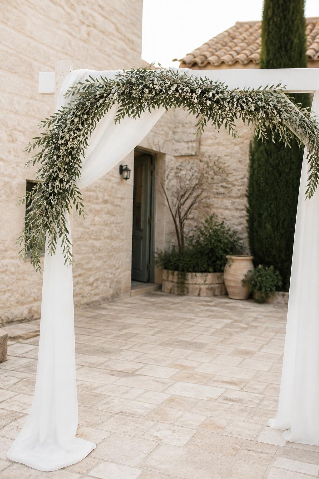 Detailed image prompt: A realistic photo of an olive branch wedding garland draped across a simple white ceremony arch, soft green olive leaves, small ivory flowers, flowing white fabric, stone courtyard wedding setting, natural Mediterranean style, no people, bright daylight.
