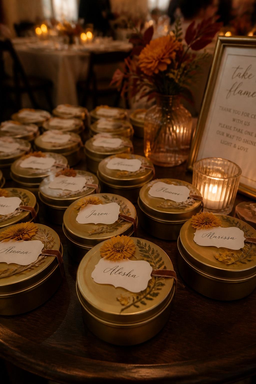 Realistic DSLR photo of small matte gold candle tins arranged on a dark wooden wedding favor table, each lid decorated with pressed marigold petals, tiny dried fern pieces, and cream name labels tied with thin velvet ribbon, warm candlelight in the background, blurred fall wedding tables behind, cozy elegant autumn atmosphere, shallow depth of field.