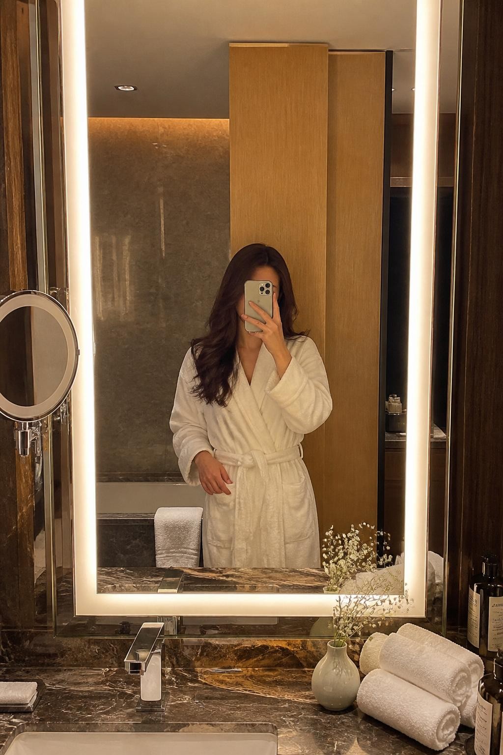 Image Prompt:
A realistic hotel bathroom mirror photoshoot of an adult woman wearing a plush white hotel robe, standing in front of a large illuminated bathroom mirror, holding a phone slightly low so her face is mostly covered. The bathroom has marble counters, rolled towels, a small vase, warm vanity lighting, skincare bottles, and a clean luxury hotel feel. The pose is relaxed and elegant, with soft reflections and realistic lighting. DSLR-style photo-realistic image, no clear facial details, vertical format. safe image