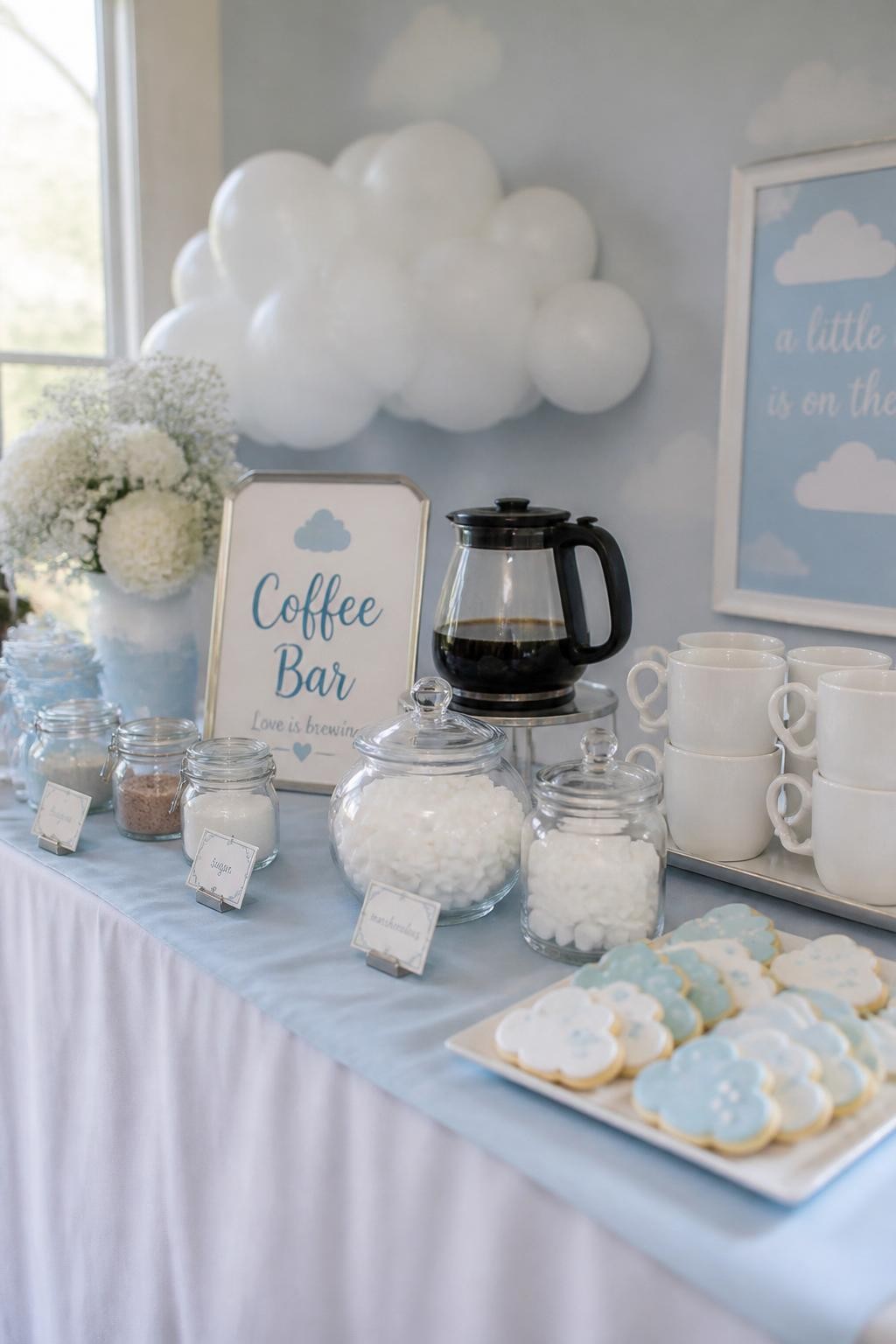 A realistic DSLR photo of a baby shower coffee bar with a soft blue cloud theme, pale blue table runner, white mugs, clear coffee carafe, glass jars of marshmallows and sugar cubes, blue and white cloud-shaped cookies on a tray, small silver labels, fluffy white balloon clouds behind the table, soft daylight, dreamy baby shower party styling, no people.
