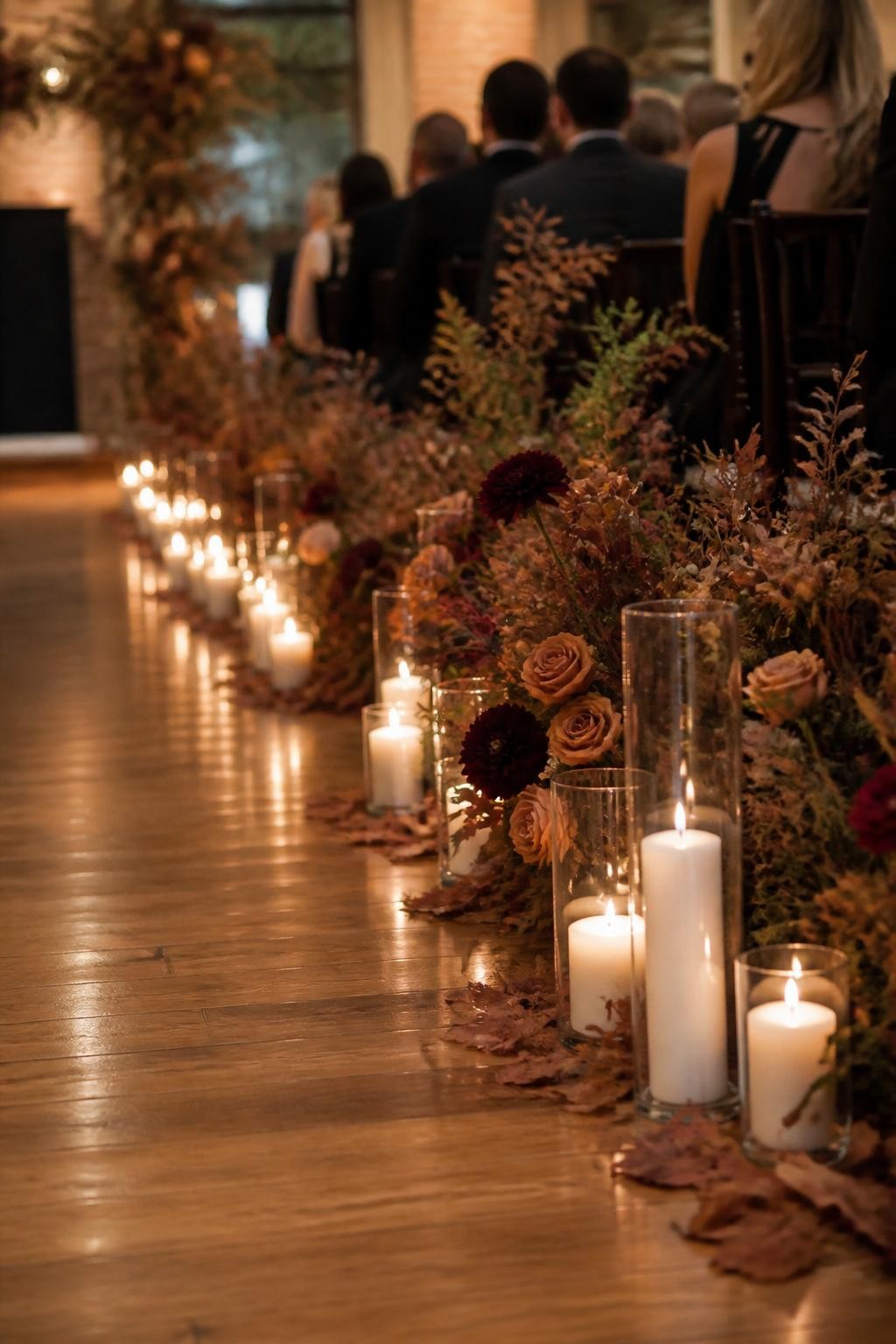 Realistic DSLR photo of an indoor fall wedding ceremony aisle lined with clusters of glass hurricane candles, ivory pillar candles, rust roses, burgundy dahlias, dried leaves, and dark greenery, polished wooden floor, warm candlelight, blurred guests seated in formal attire, romantic moody wedding atmosphere.