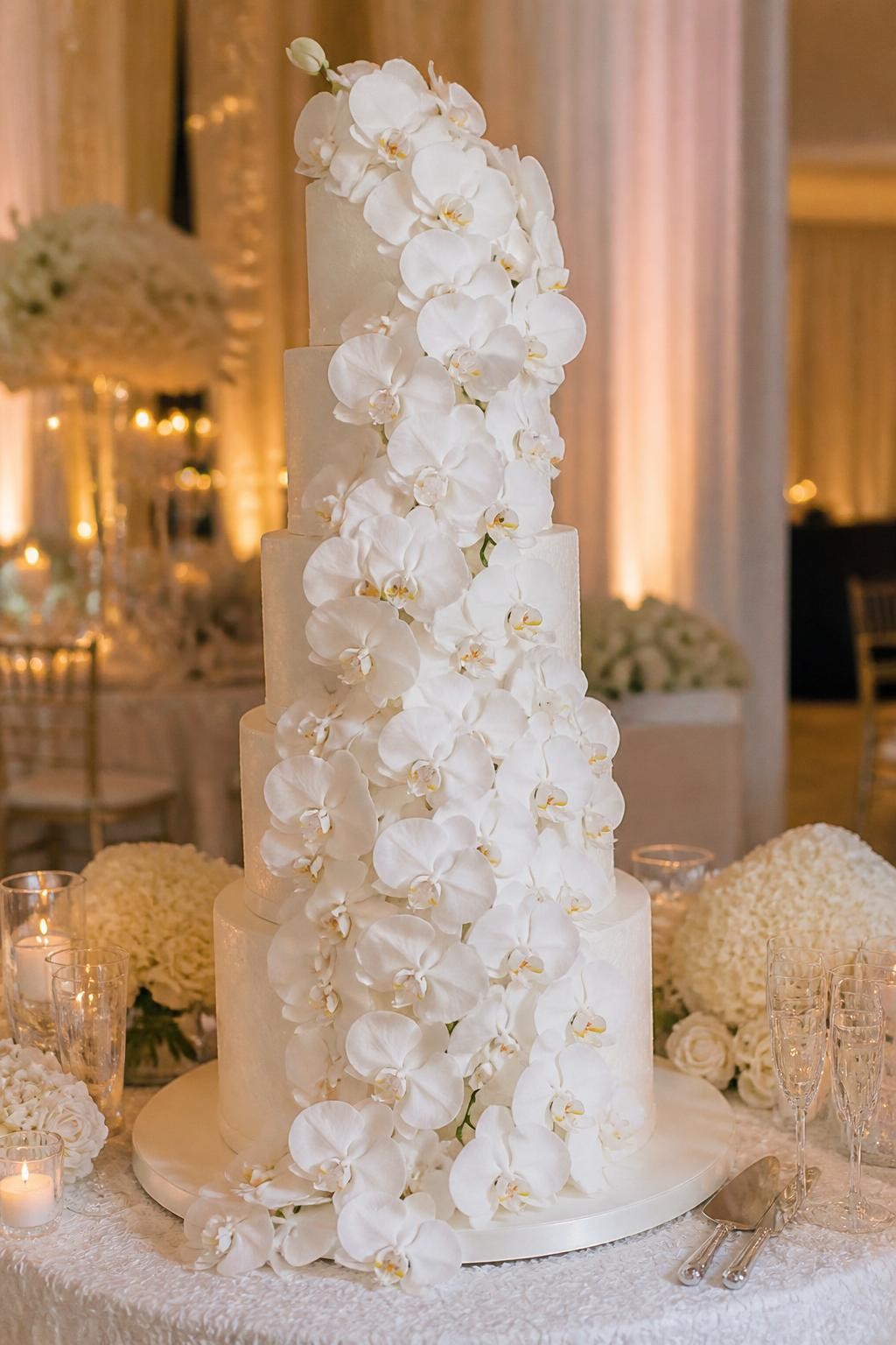 Image Prompt: Ultra-realistic wedding party photo of an extravagant wedding cake with a dramatic waterfall of white orchids cascading down tall tiers. Style the cake on a luxury reception cake table with candles, soft floral decor, elegant glassware, and a beautiful wedding venue background. Keep the cake front and center. No visible faces. If guests appear, show only side views in blur, back views, cropped torsos, or hands near the cake table. Realistic orchid petals, polished icing finish, romantic soft light, DSLR wedding editorial photography. (no watermarks on images)