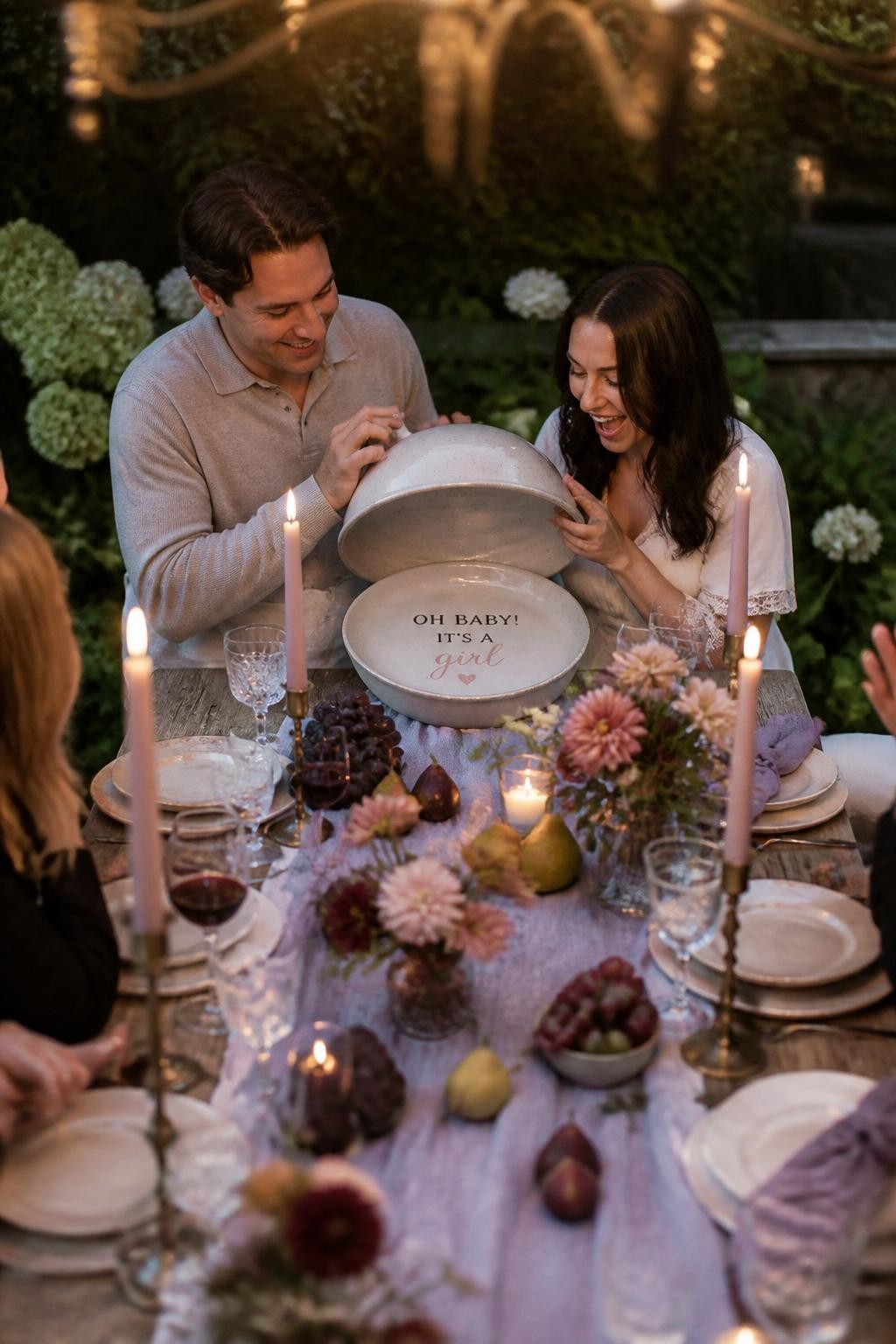 A realistic editorial photo of a September gender reveal garden dinner, long outdoor table with linen runner, taper candles, dahlias, pears, figs, grapes, ceramic plates, parents lifting a covered dish to reveal a baby message and soft color detail, warm evening light, elegant dinner party photography