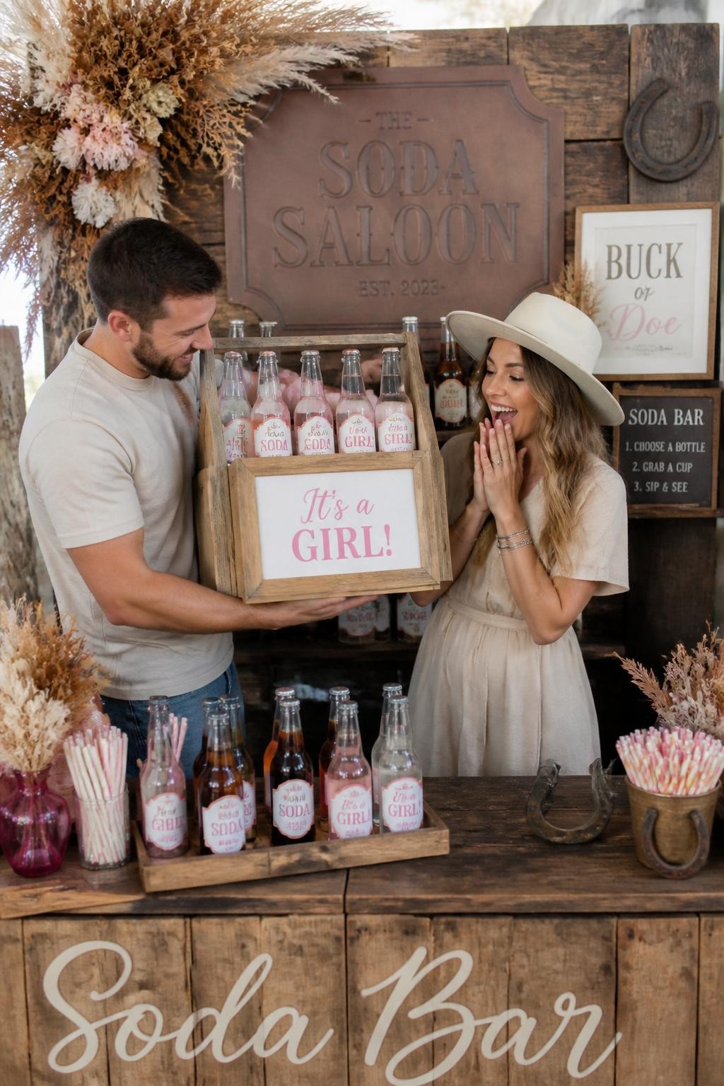 A realistic editorial photo of a fun modern western gender reveal soda bar, rustic wood counter with glass bottles, custom soda labels, striped paper straws, leather sign reading saloon, dried flowers, parents opening one special bottle or lifting a bottle crate to reveal the baby message, bright cheerful party styling