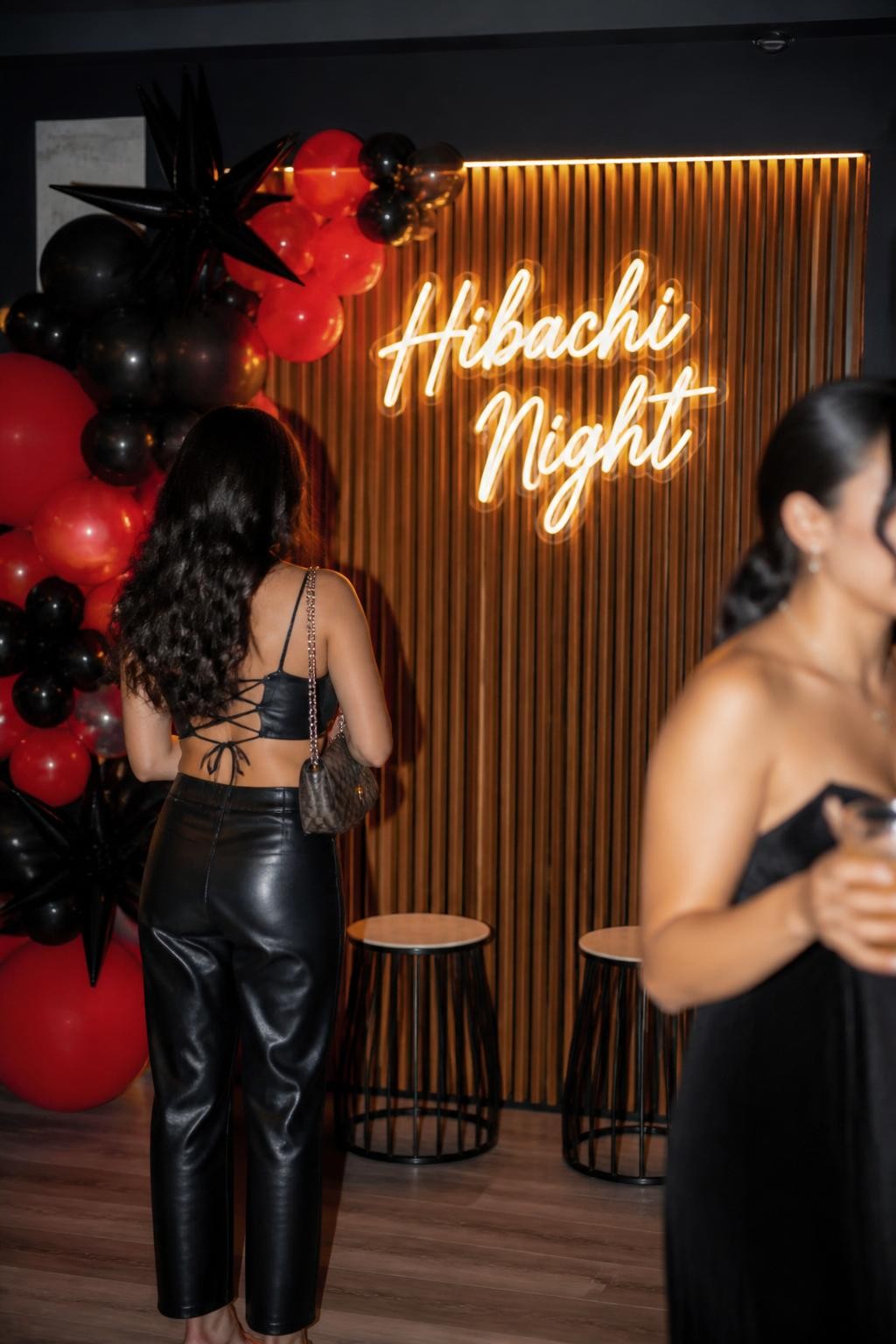 Image Prompt:
A realistic DSLR photo of a modern party photo corner with a glowing “Hibachi Night” neon sign, wood slat backdrop, a cluster of black and red balloons, small stools, and one guest standing with back to camera while another is softly blurred, fun but stylish party setting, DSLR flash photography look.
