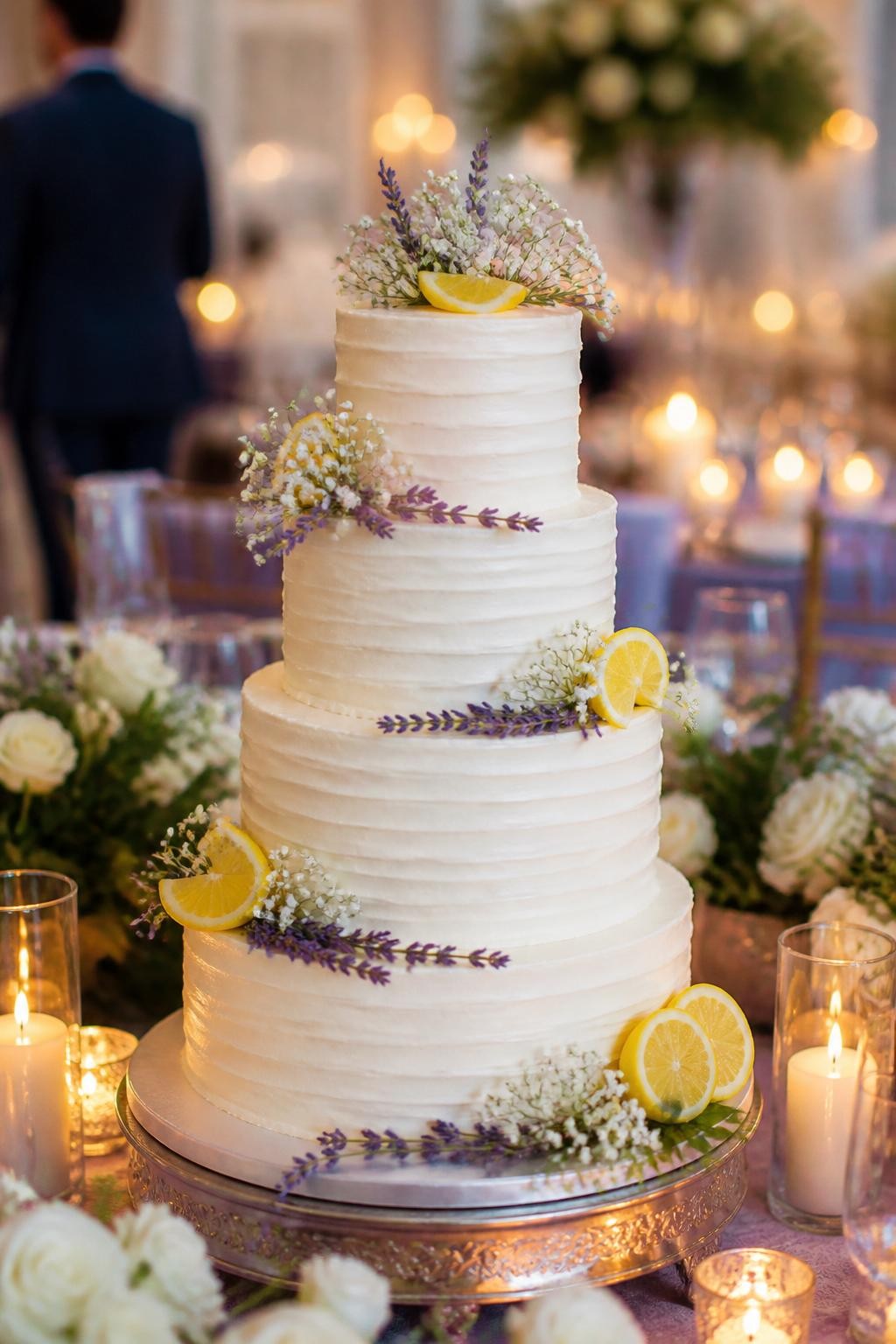 Image Prompt: Ultra-realistic wedding party photo of a lemon and lavender wedding cake with smooth white or pale cream tiers, fresh lemon accents, lavender sprigs, and small white flowers arranged beautifully on the cake. Style the cake on a reception cake table with candles, lavender and white floral decor, fine linens, glassware, and a softly blurred wedding venue backdrop. Keep the cake as the main focus. No visible faces. If guests appear, show only back views, side views in blur, cropped hands, or neck-down figures. Realistic lavender texture, fresh lemon detail, romantic DSLR wedding photography. (no watermarks on images)