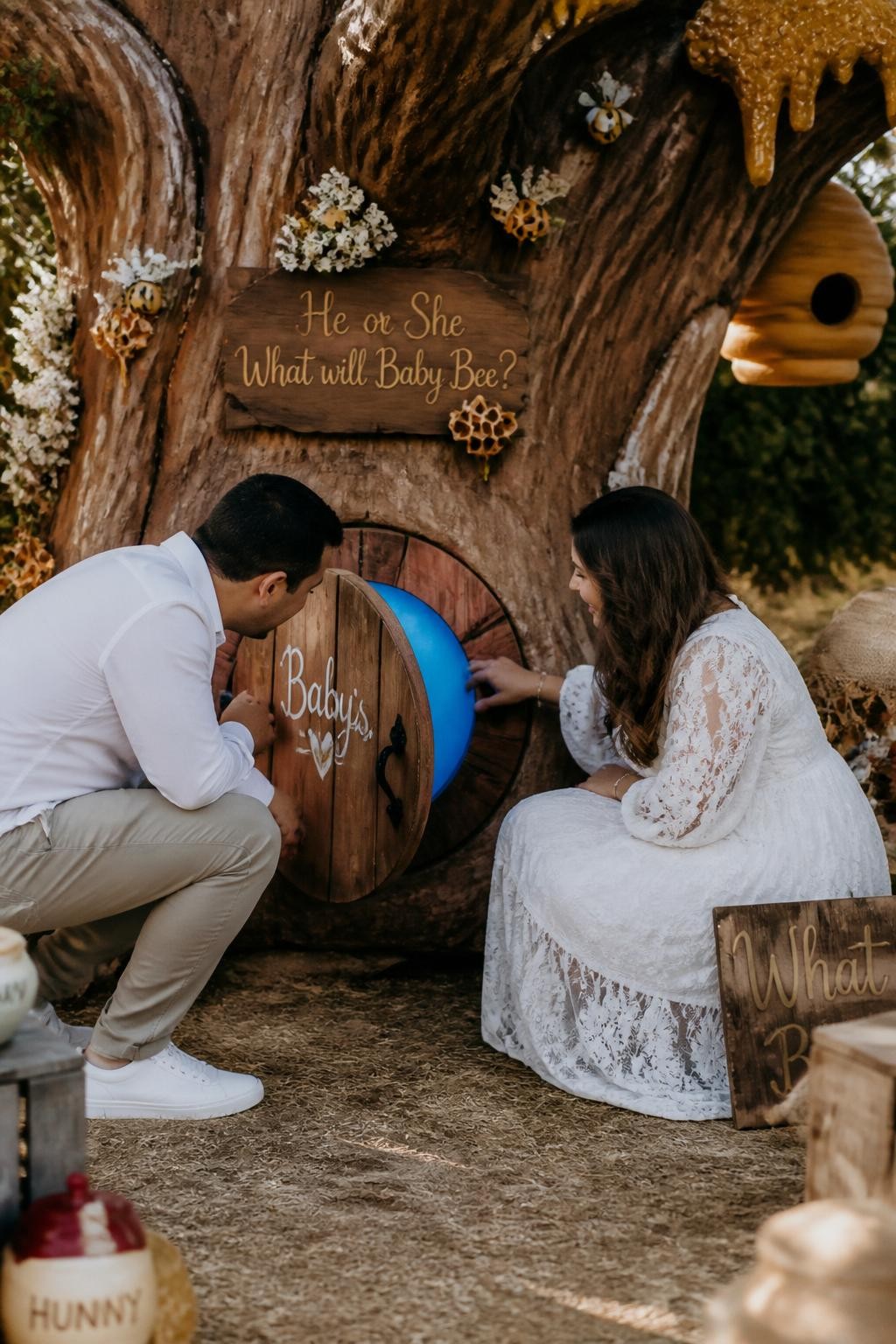A realistic editorial photo of a gender reveal party backdrop styled like a soft storybook honey tree, warm brown tree trunk, golden honey details, tiny bees, parents opening a small round tree door to reveal one glowing color inside, rustic woodland party setting, vertical DSLR lifestyle photo