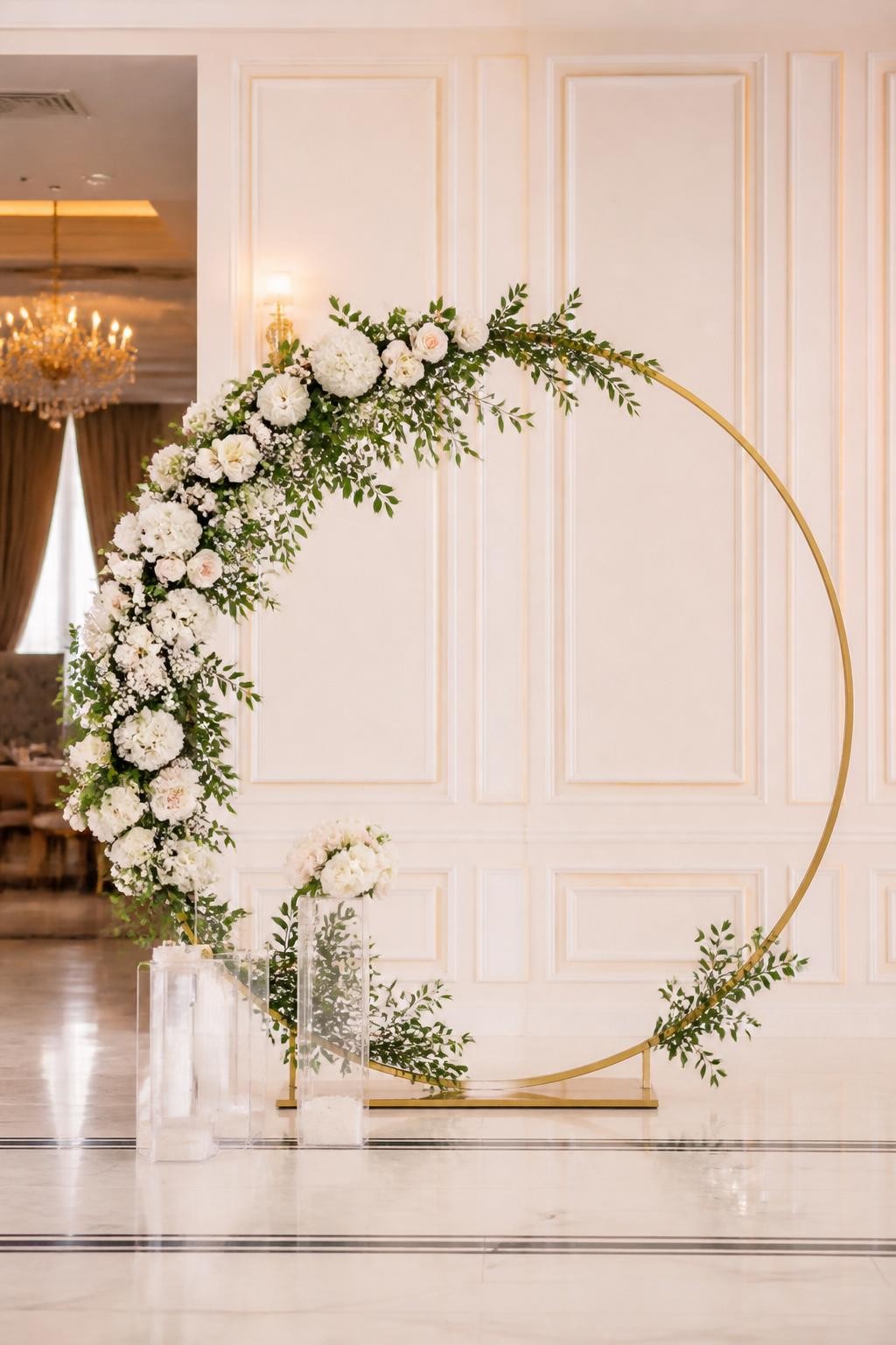 Image Prompt:
A single realistic editorial-style photo of a floral hoop backdrop with a large gold circular metal frame decorated halfway with white roses, blush flowers, baby’s breath, and greenery, clear plinths at the base, elegant indoor celebration space, no people, no faces, warm soft lighting, realistic event photography, no text, no watermark.