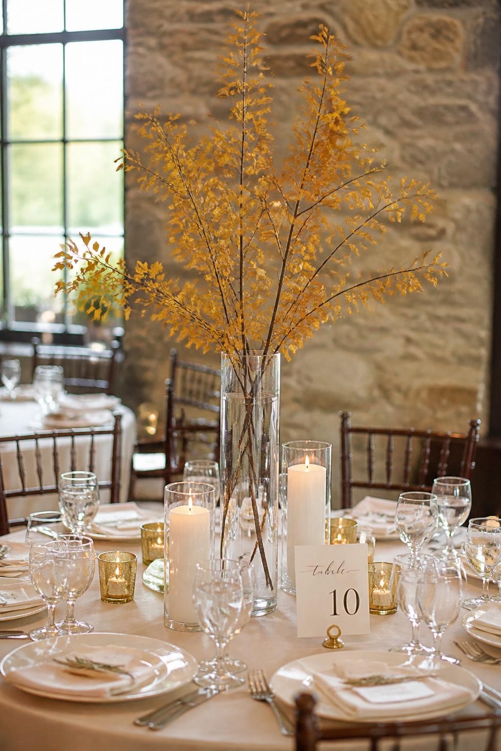 Realistic DSLR photo of a simple fall wedding centerpiece with a tall clear glass vase holding delicate autumn branches with golden leaves, placed in the center of a round reception table with ivory candles, cream plates, and soft neutral linens, elegant natural indoor light.