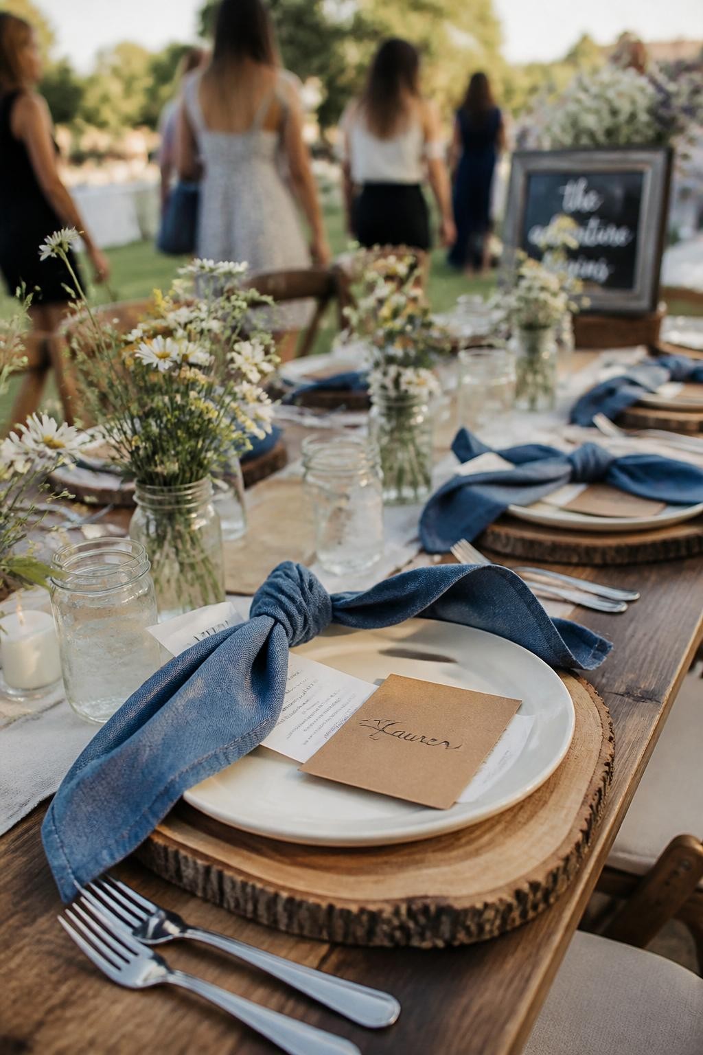 A realistic DSLR photo of a rustic graduation party table with denim-blue napkins, wildflower jars, wooden chargers, cream plates, kraft paper place cards, soft yellow flowers, and a few guests walking in the background with faces not visible and softly blurred, sunny outdoor lighting, modern rustic party styling.