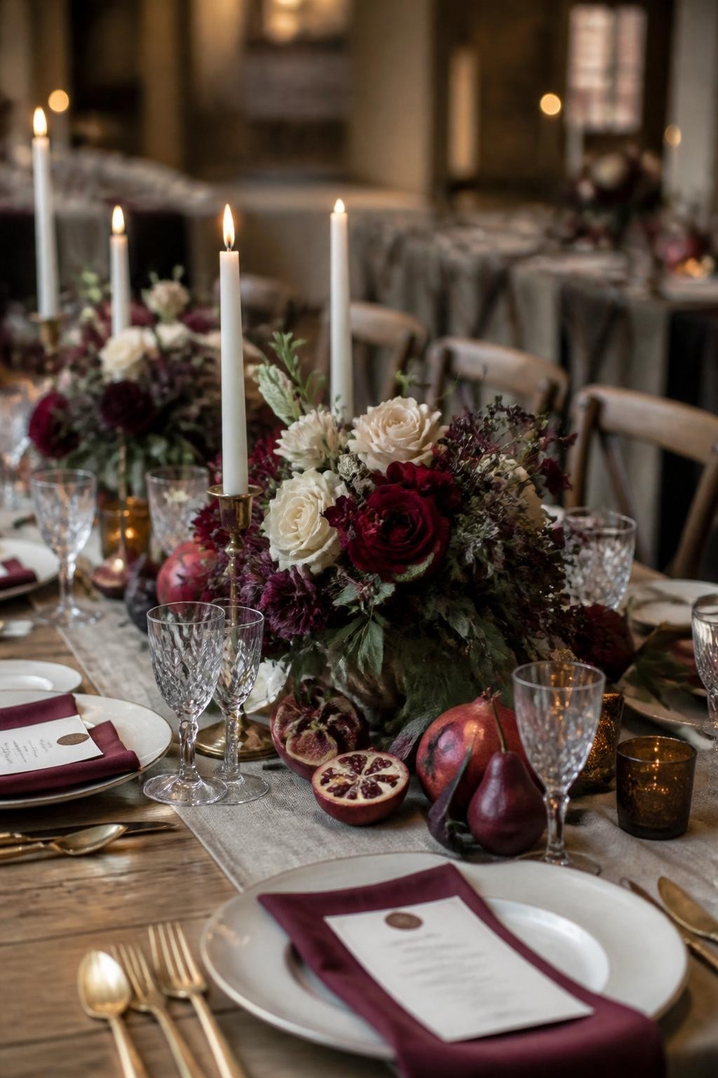 Realistic DSLR photo of a late fall wedding tablescape with fresh pomegranates, dark red pears, figs, cream plates, gold flatware, burgundy napkins, ivory taper candles, and low floral arrangements in deep red, plum, and cream, warm candlelit reception setting.