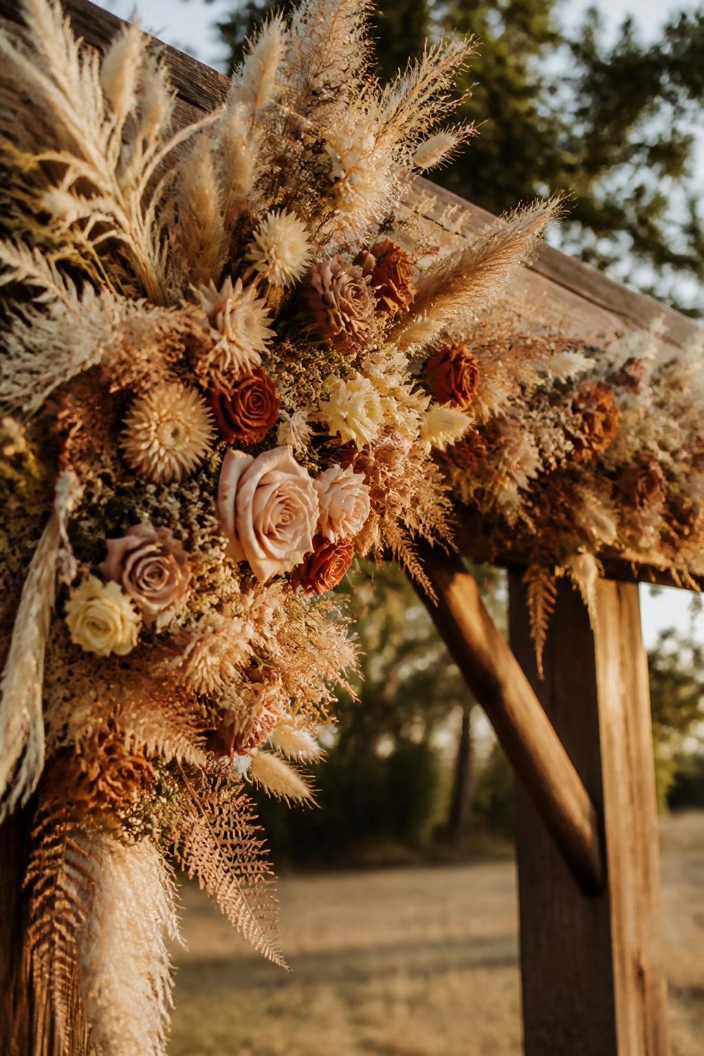 Detailed image prompt: A realistic DSLR-style photo of a dried flower wedding garland draped along a rustic wooden ceremony arch, dried roses, bunny tails, strawflowers, pampas grass, preserved ferns, beige, cream, rust, blush, and muted orange tones, outdoor boho wedding setup, no people, golden hour light.