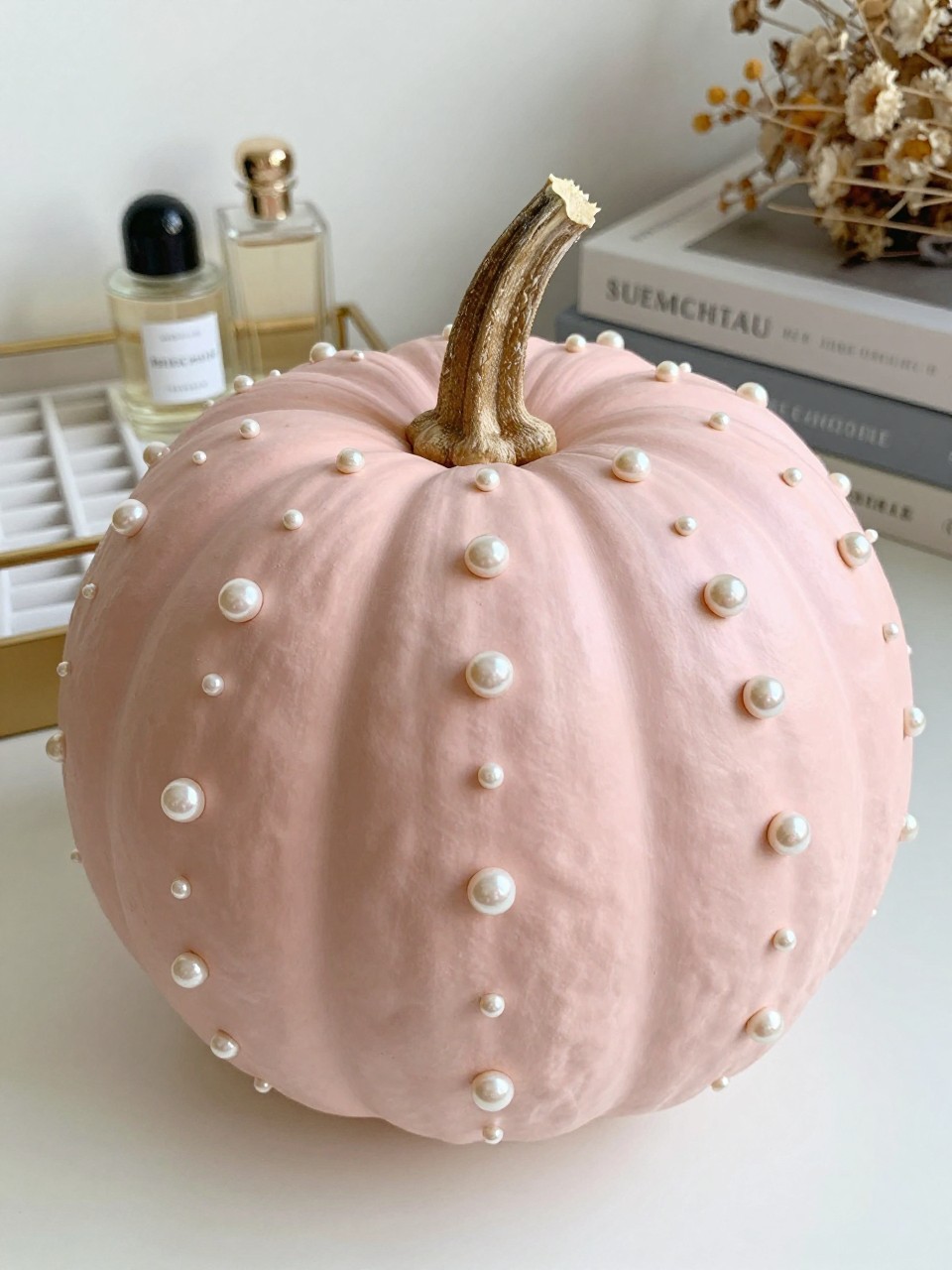 Horizontal 16:9 casual iPhone camera photo of a matte pale-pink real pumpkin decorated with small hand-painted pearl-like dots in warm ivory and soft champagne, dots following selected natural ridges in loose vertical rows, natural brown stem, displayed on a dressing table beside a jewelry tray, perfume bottles, neutral books, and dried flowers, soft morning daylight, subtle pearlescent sheen, no glued plastic pearls, no text.
