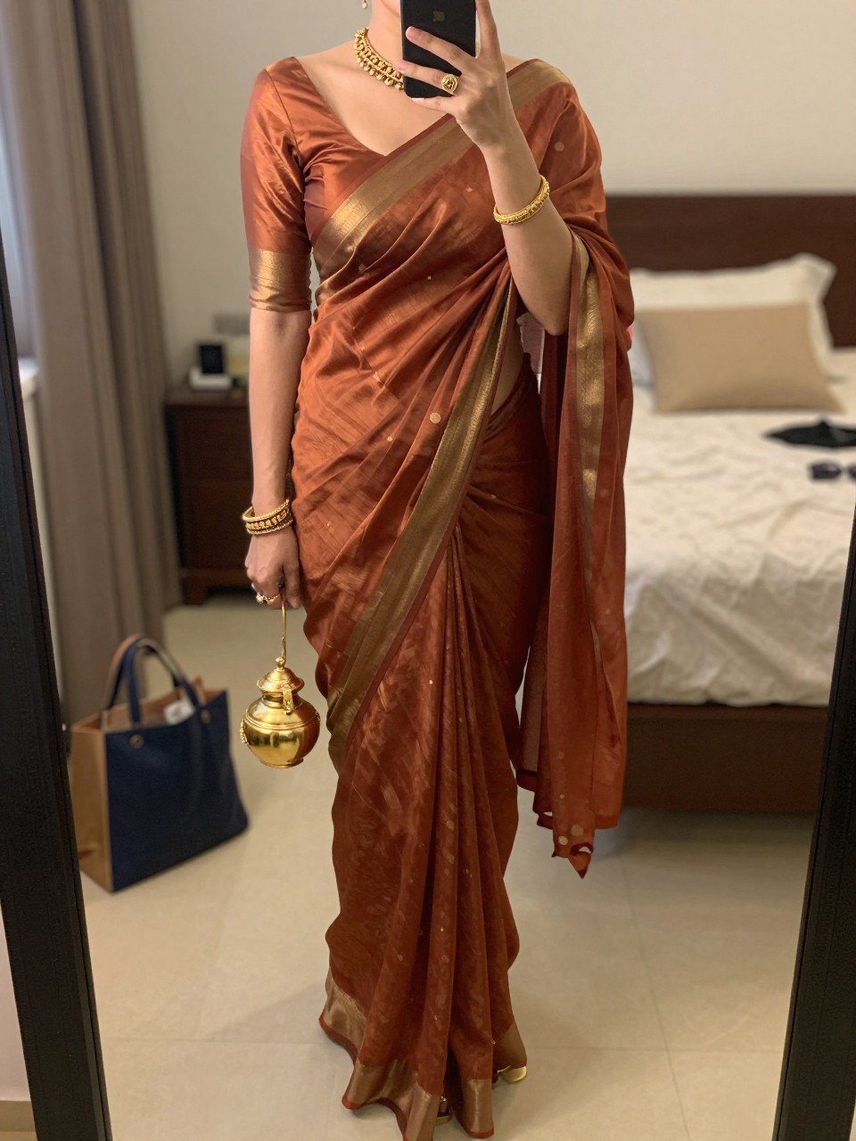 Image prompt
mirror selfie of a woman wearing a rust silk blend saree with a subtle sheen and a matching blouse, antique gold jewelry set, gold sandals, and a small gold potli bag, holding a phone in front of a mirror, cropped at NECK so face doesn't show, with a natural casual selfie pose, in a feminine-themed bedroom, warm moody lighting, including small lived-in details like a tote bag on the floor, sunglasses on a dresser, or soft bedding in the background, latest iPhone photo quality.
