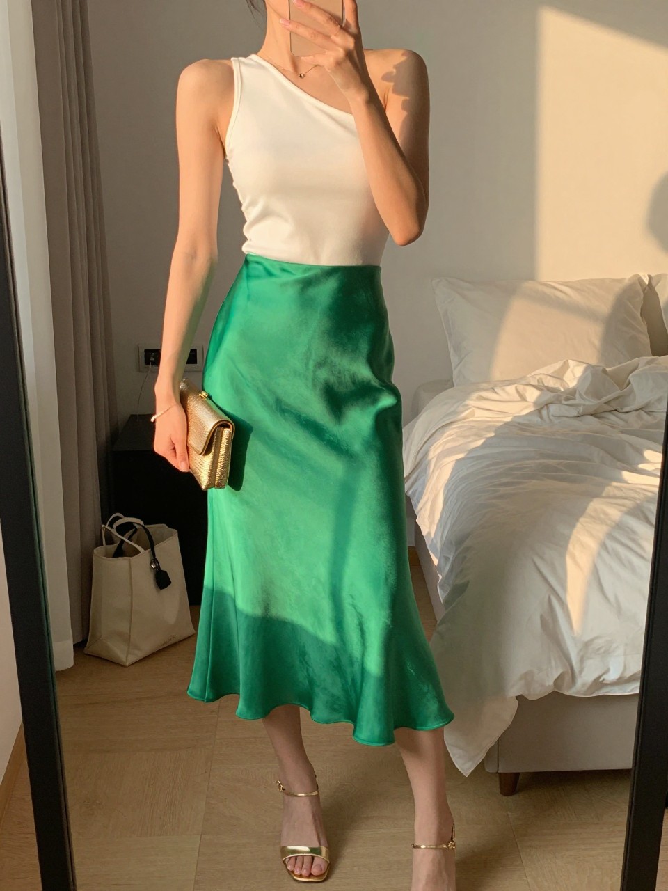 Image prompt
mirror selfie of a woman wearing a bright green satin midi skirt with a fitted white one-shoulder top, metallic gold heeled sandals, and a gold clutch, holding a phone in front of a mirror, cropped at NECK so face doesn't show, with a natural casual selfie pose, in a feminine-themed bedroom, evening golden hour, including small lived-in details like a tote bag on the floor and soft bedding in the background, latest iPhone photo quality.