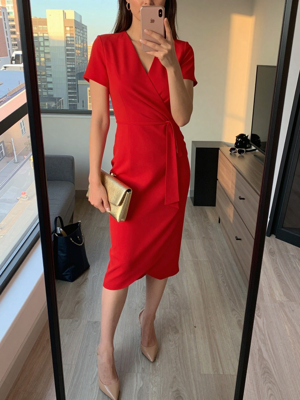 Image prompt
mirror selfie of a woman wearing a bright red wrap dress with nude pointed-toe heels, a gold clutch, and gold hoop earrings, holding a phone in front of a mirror, cropped at NECK so face doesn't show, with a natural casual selfie pose, in a city apartment, evening golden hour, including small lived-in details like a tote bag on the floor and sunglasses on a dresser, latest iPhone photo quality.