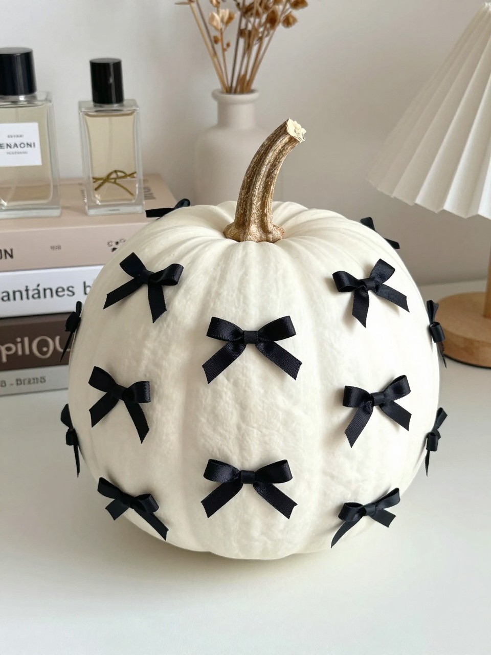 Horizontal 16:9 casual iPhone photo of a matte white real pumpkin decorated with scattered tiny hand-painted black bows, bows slightly varied in size and direction with short ribbon tails, natural twisted stem, displayed on a vanity table beside perfume bottles, neutral books, dried flowers, and a small ceramic lamp, gentle morning daylight, cute but polished handmade details, no text.