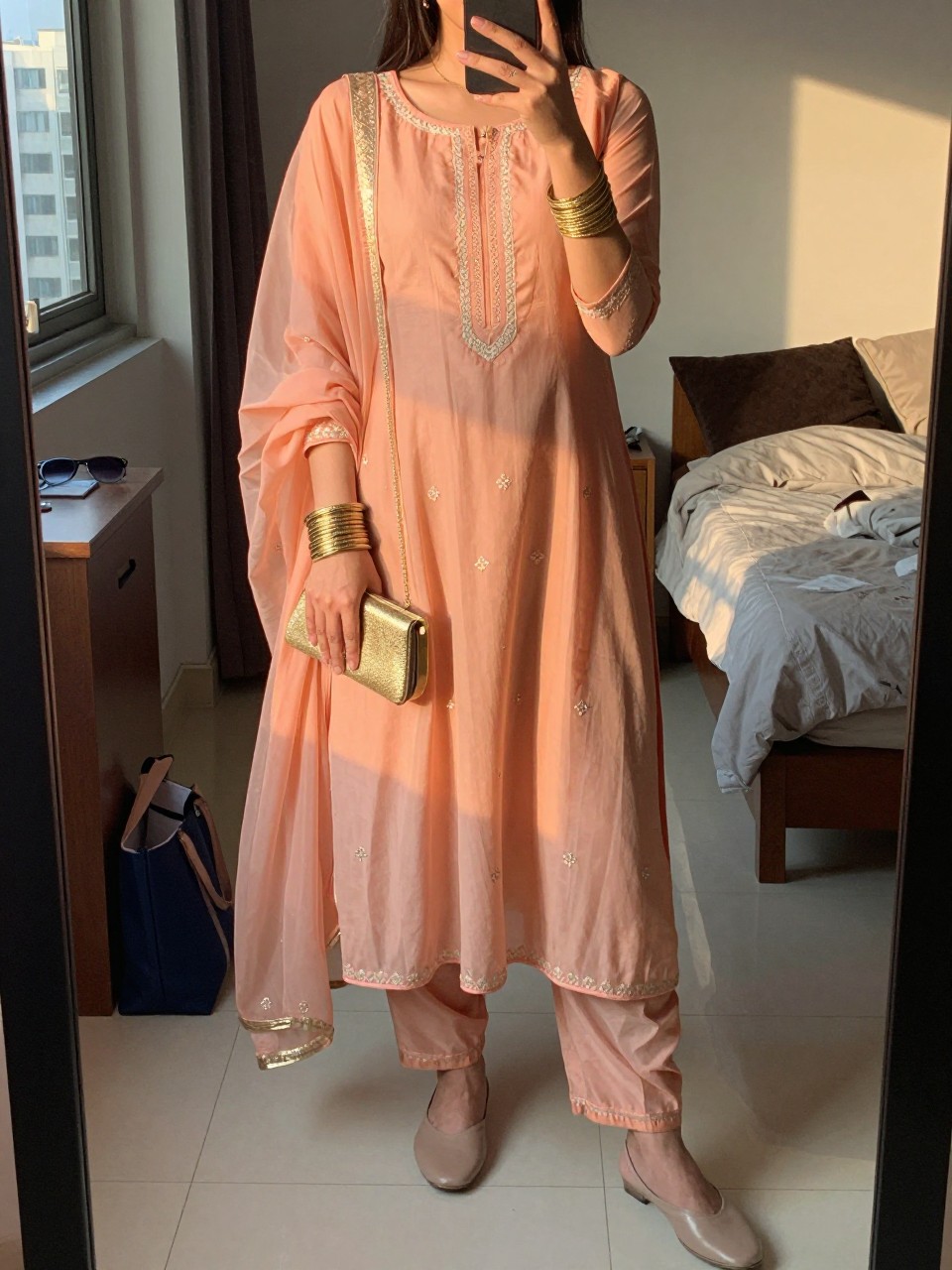 Image prompt
mirror selfie of a woman wearing a peach chiffon anarkali suit with light embroidery and a sheer matching dupatta, stacked gold bangles, nude juttis, and a small gold clutch, holding a phone in front of a mirror, cropped at NECK so face doesn't show, with a natural casual selfie pose, in a city apartment, evening golden hour, including small lived-in details like a tote bag on the floor, sunglasses on a dresser, or soft bedding in the background, latest iPhone photo quality.