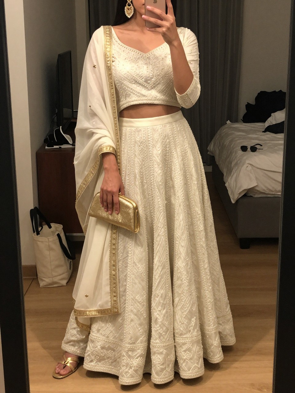 Image prompt
mirror selfie of a woman wearing an ivory lehenga with delicate threadwork and an ivory dupatta with a gold border, gold chandelier earrings, gold strappy heels, and a small gold clutch, holding a phone in front of a mirror, cropped at NECK so face doesn't show, with a natural casual selfie pose, in a city apartment, warm moody lighting, including small lived-in details like a tote bag on the floor, sunglasses on a dresser, or soft bedding in the background, latest iPhone photo quality.