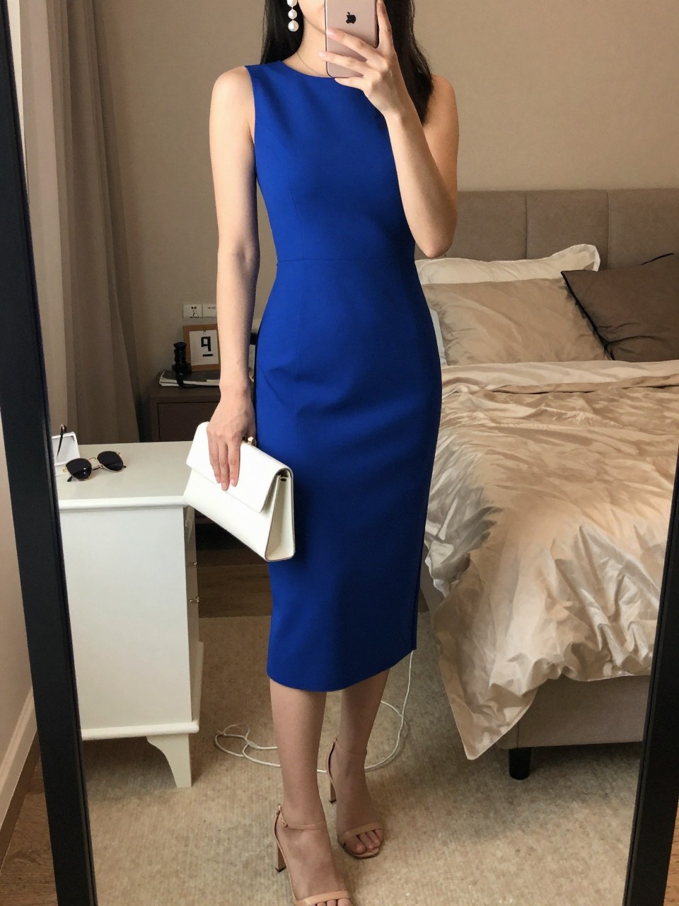 Image prompt
mirror selfie of a woman wearing a cobalt blue midi dress with nude heeled sandals, pearl drop earrings, and a white clutch, holding a phone in front of a mirror, cropped at NECK so face doesn't show, with a natural casual selfie pose, in a feminine-themed bedroom, warm moody lighting, including small lived-in details like soft bedding and sunglasses on a dresser, latest iPhone photo quality.
