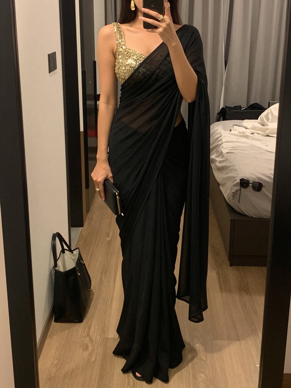 Image prompt
mirror selfie of a woman wearing a black georgette saree with a clean drape and a gold sequin blouse, gold drop earrings, black heels, and a small black clutch, holding a phone in front of a mirror, cropped at NECK so face doesn't show, with a natural casual selfie pose, in a city apartment, warm moody lighting, including small lived-in details like a tote bag on the floor, sunglasses on a dresser, or soft bedding in the background, latest iPhone photo quality.