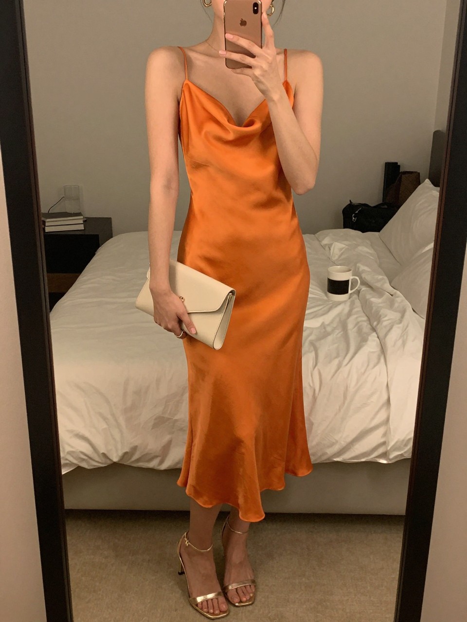 Image prompt
mirror selfie of a woman wearing an orange satin midi dress with metallic champagne heeled sandals, a cream clutch, and small gold hoop earrings, holding a phone in front of a mirror, cropped at NECK so face doesn't show, with a natural casual selfie pose, in a feminine-themed bedroom, warm moody lighting, including small lived-in details like soft bedding and a coffee mug nearby, latest iPhone photo quality.
