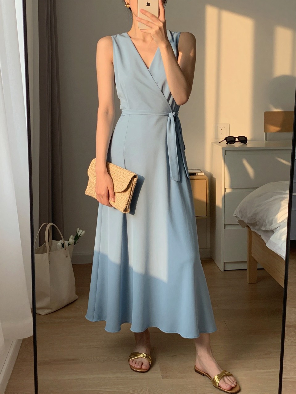 Image prompt
mirror selfie of a woman wearing a soft blue wrap maxi dress with a flowy skirt, gold flat sandals with a dressy finish, small gold drop earrings, and a small raffia clutch, holding a phone in front of a mirror, cropped at NECK so face doesn't show, with a natural casual selfie pose, in a city apartment, evening golden hour, including small lived-in details like a tote bag on the floor, sunglasses on a dresser, or soft bedding in the background, latest iPhone photo quality.