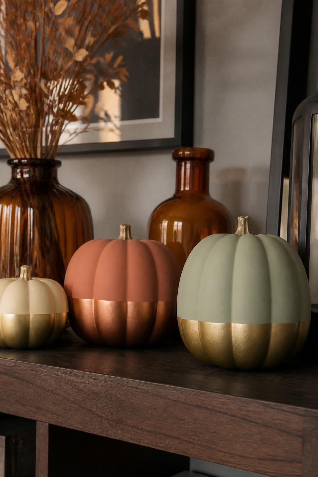 Image Prompt: A realistic DSLR photo of matte foam pumpkins in cream, terracotta, and sage green with metallic gold and copper dipped bottoms, arranged on a dark wood shelf with amber glass vases, dried branches, and soft evening light, modern stylish autumn decor, clean painted edges and realistic texture.